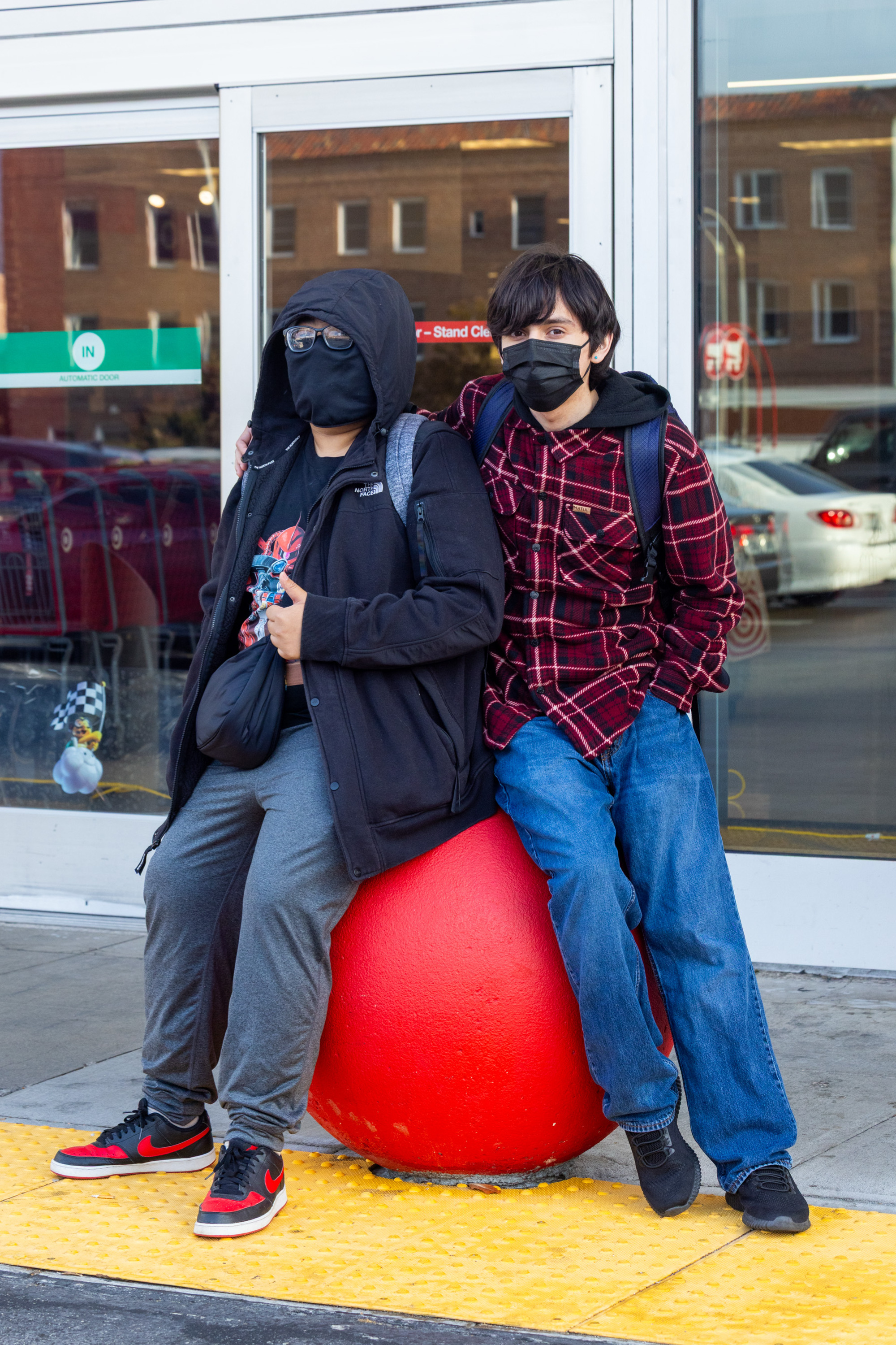 Two people, wearing masks and casual outfits, lean against a large red sphere outside a store. Both look relaxed and are posing close together.