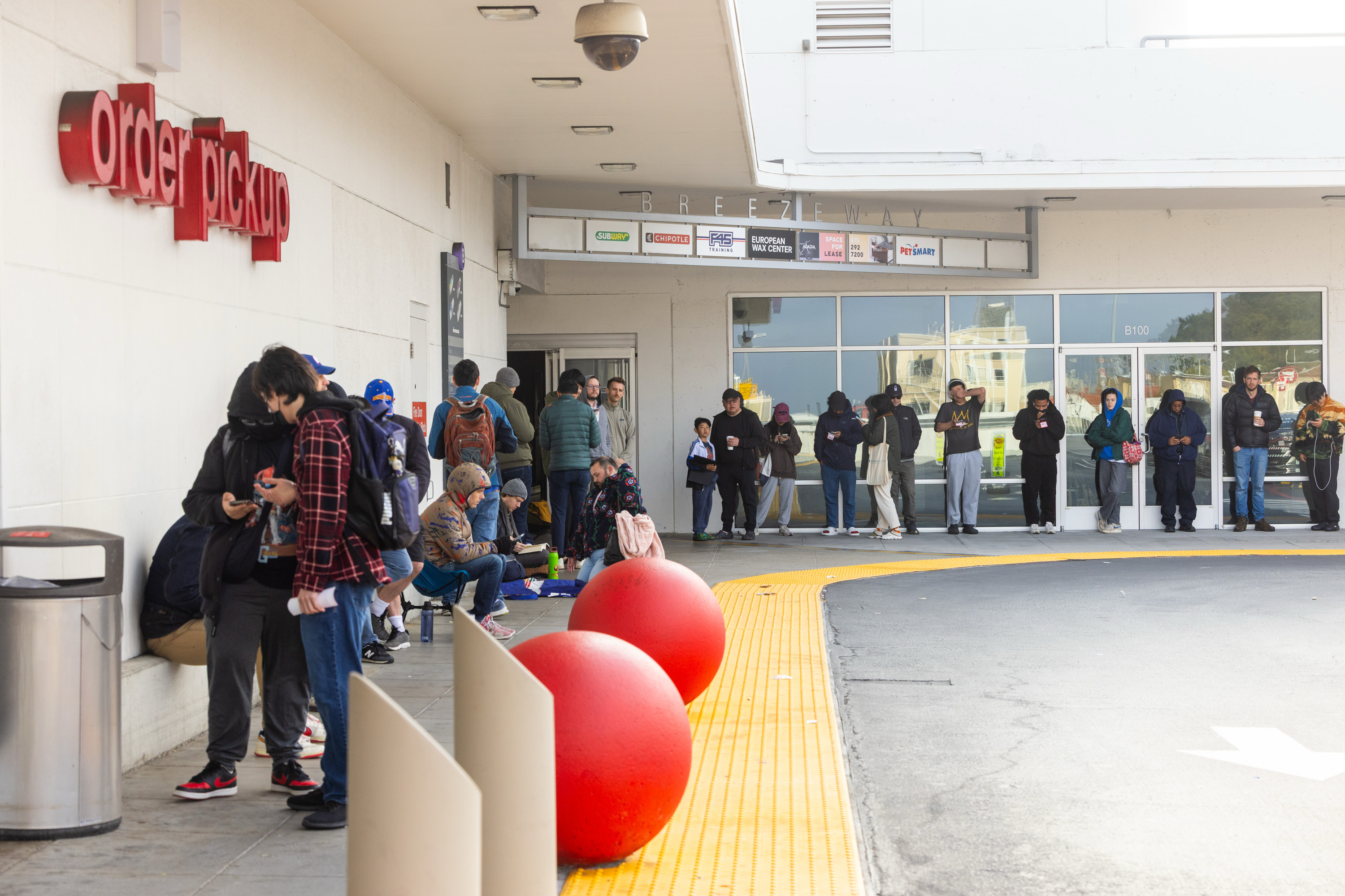 A group of people are lined up outside an "order pickup" area, some standing and others sitting, surrounded by large red spheres and a trash can.