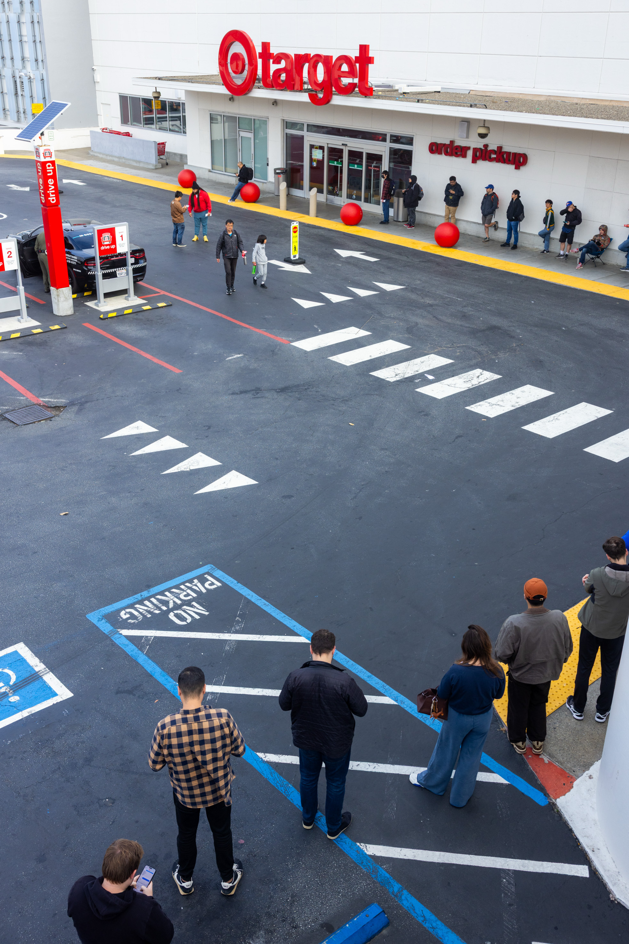 People wait in a line outside a Target store near the &quot;order pickup&quot; area, some using phones. The scene shows parking spots and red spheres decorating the entrance.