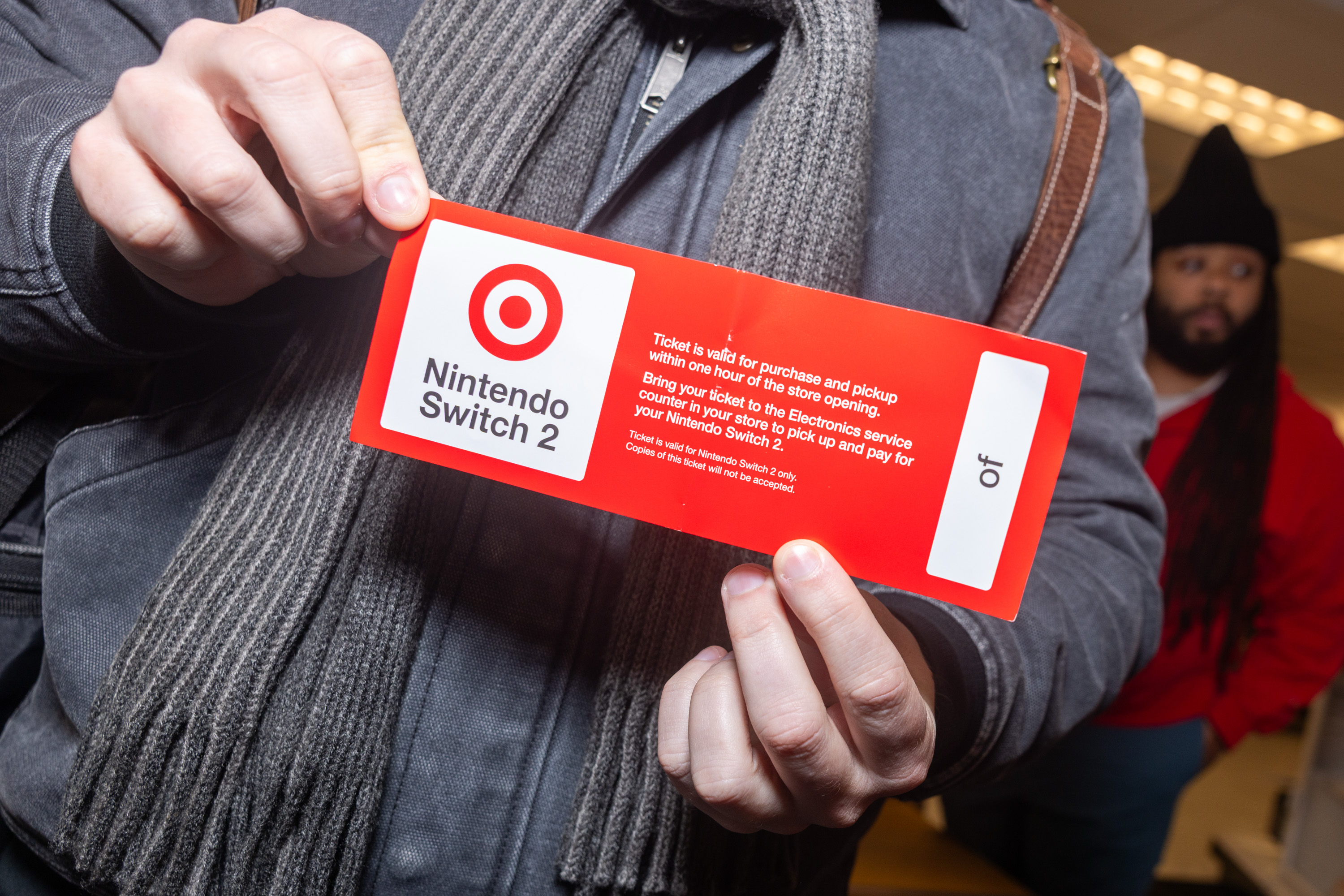 A person holds a red card with a Target logo, labeled &quot;Nintendo Switch 2.&quot; The text details a purchase and pickup process. Another person is in the background.
