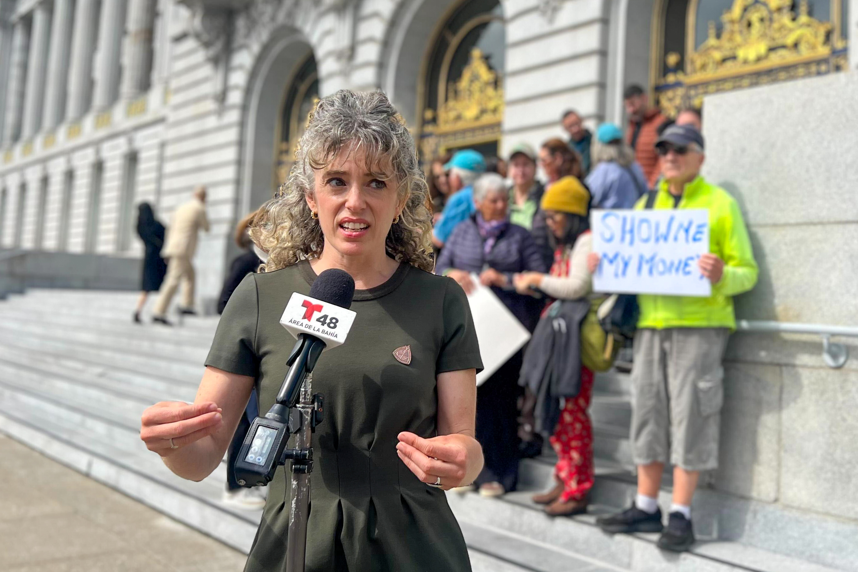 A woman stands in front of a microphone on the steps of a building. A group behind her holds a sign reading “Show me my money.” The building has ornate doors.