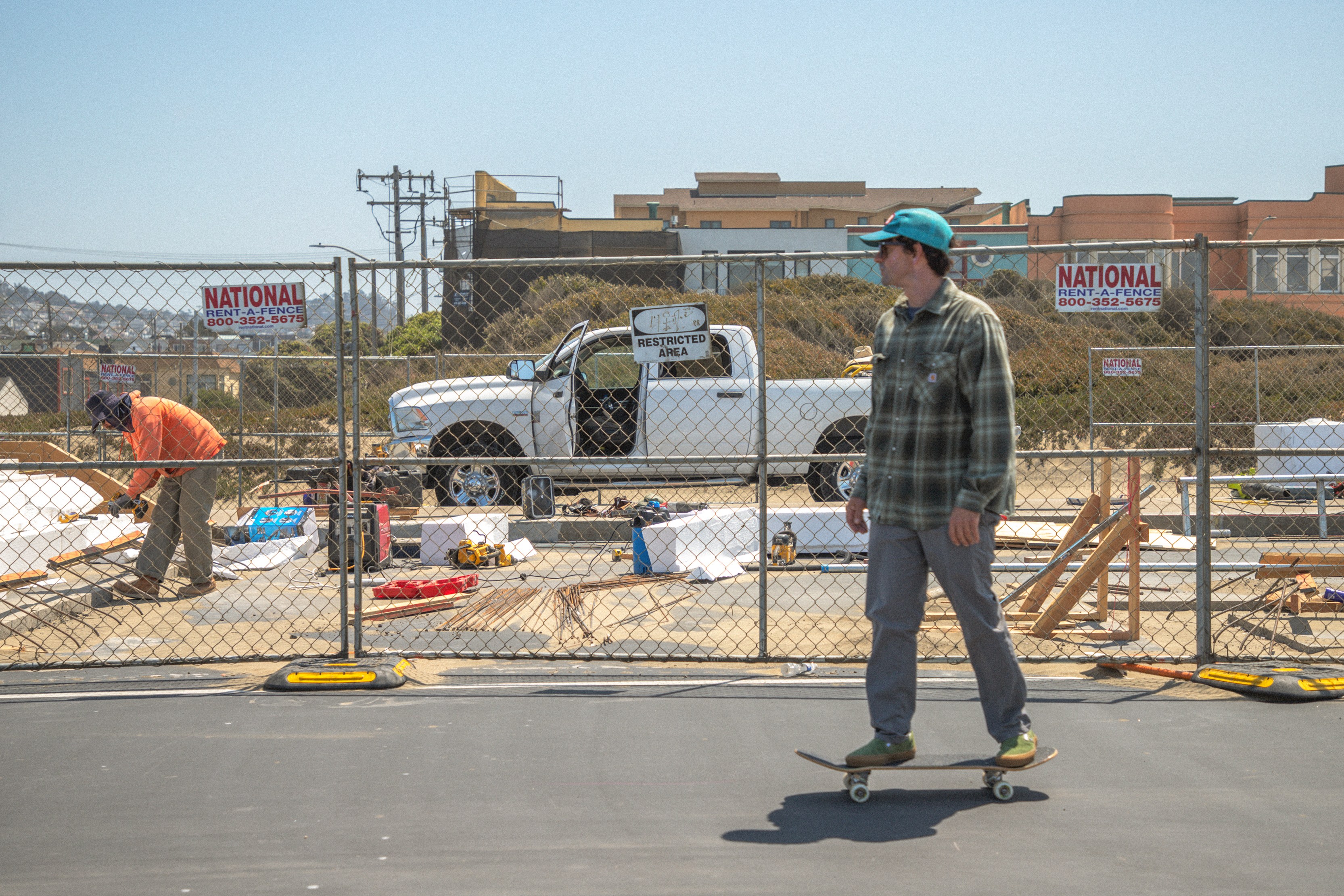 A man skateboards past a construction site, where another person is working near a pickup truck. A fence with "Restricted Area" signs surrounds the site.