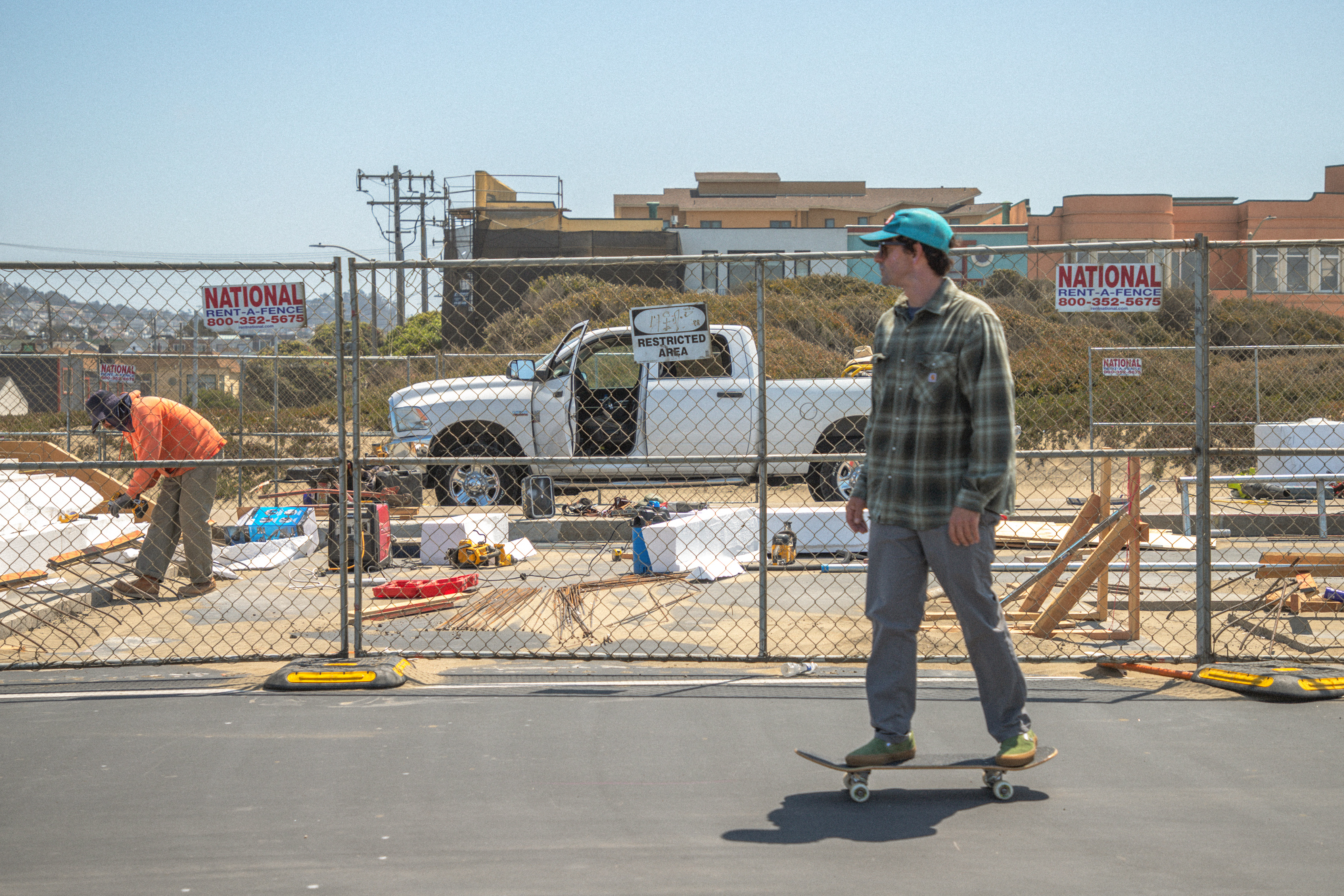 A man skateboards past a construction site, where another person is working near a pickup truck. A fence with "Restricted Area" signs surrounds the site.