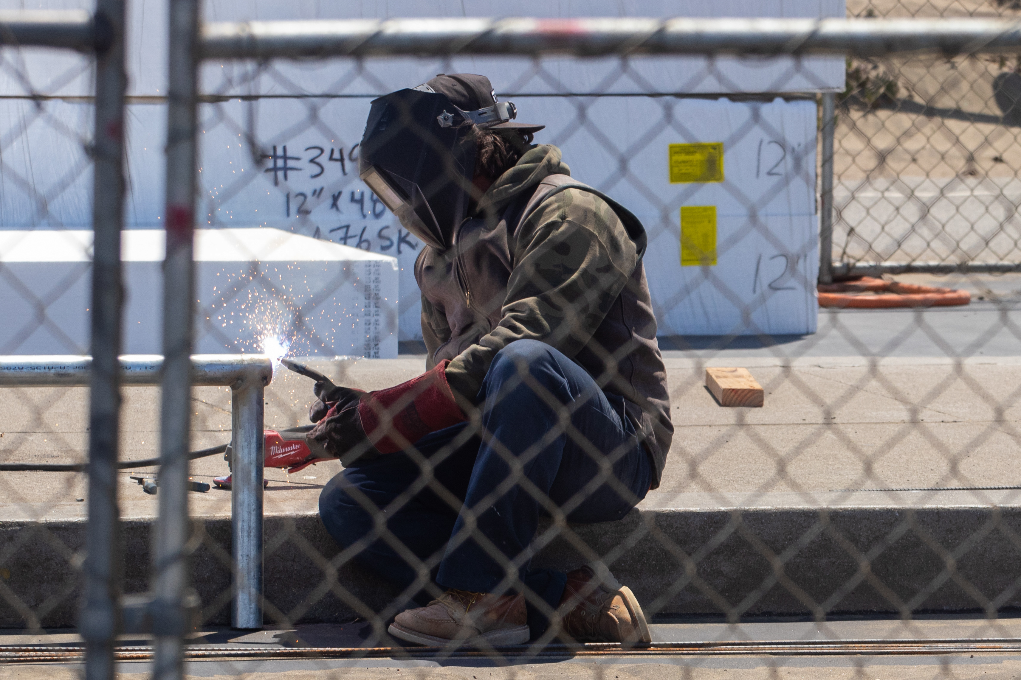 A person wearing protective gear is crouched down, welding a metal pipe, with sparks flying. They are working behind a chain-link fence.