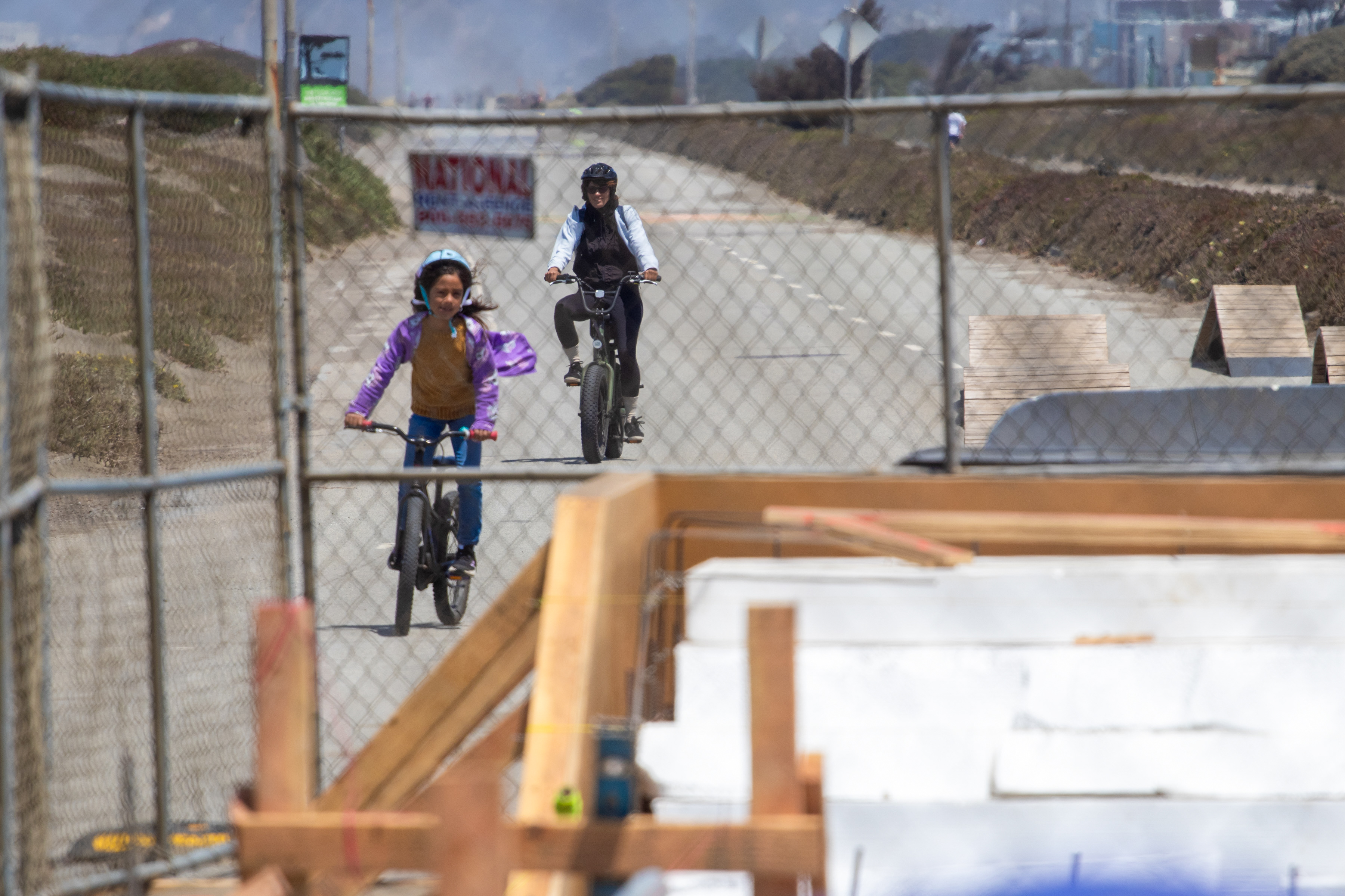 A child and an adult ride bikes on a paved road, behind a chain-link fence. There are wooden ramps on the road and a hillside flanks the scene.