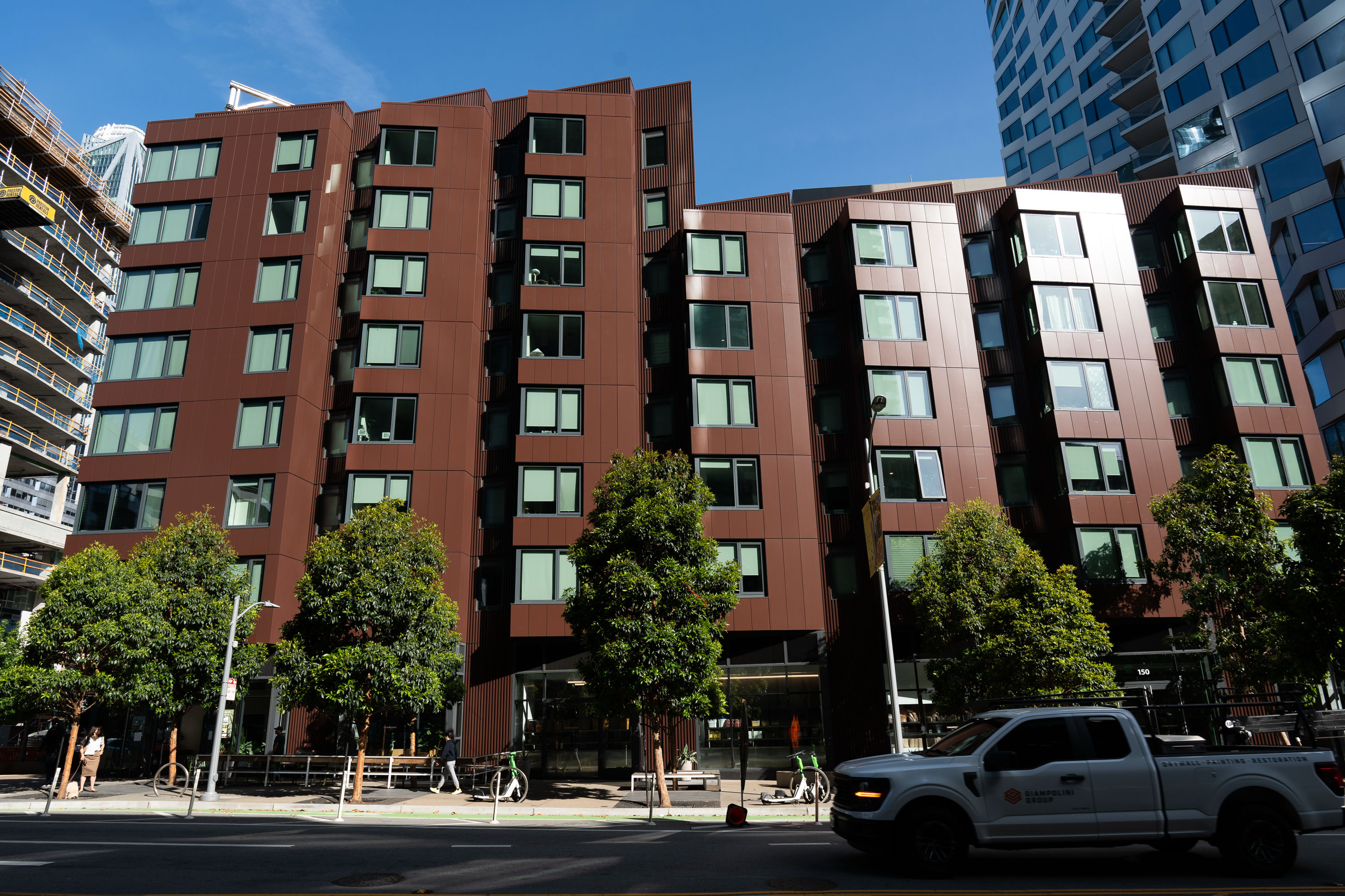 The image shows a modern, angular brown building with many windows, surrounded by trees. A white truck is driving along the street in front.