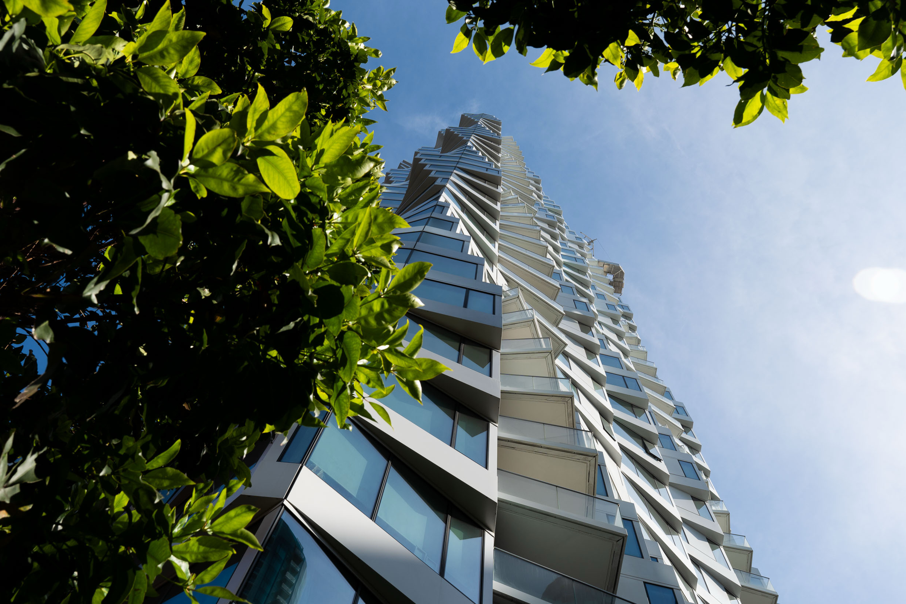 A modern, twisting skyscraper towers upward against a blue sky, framed by green leaves in the foreground.