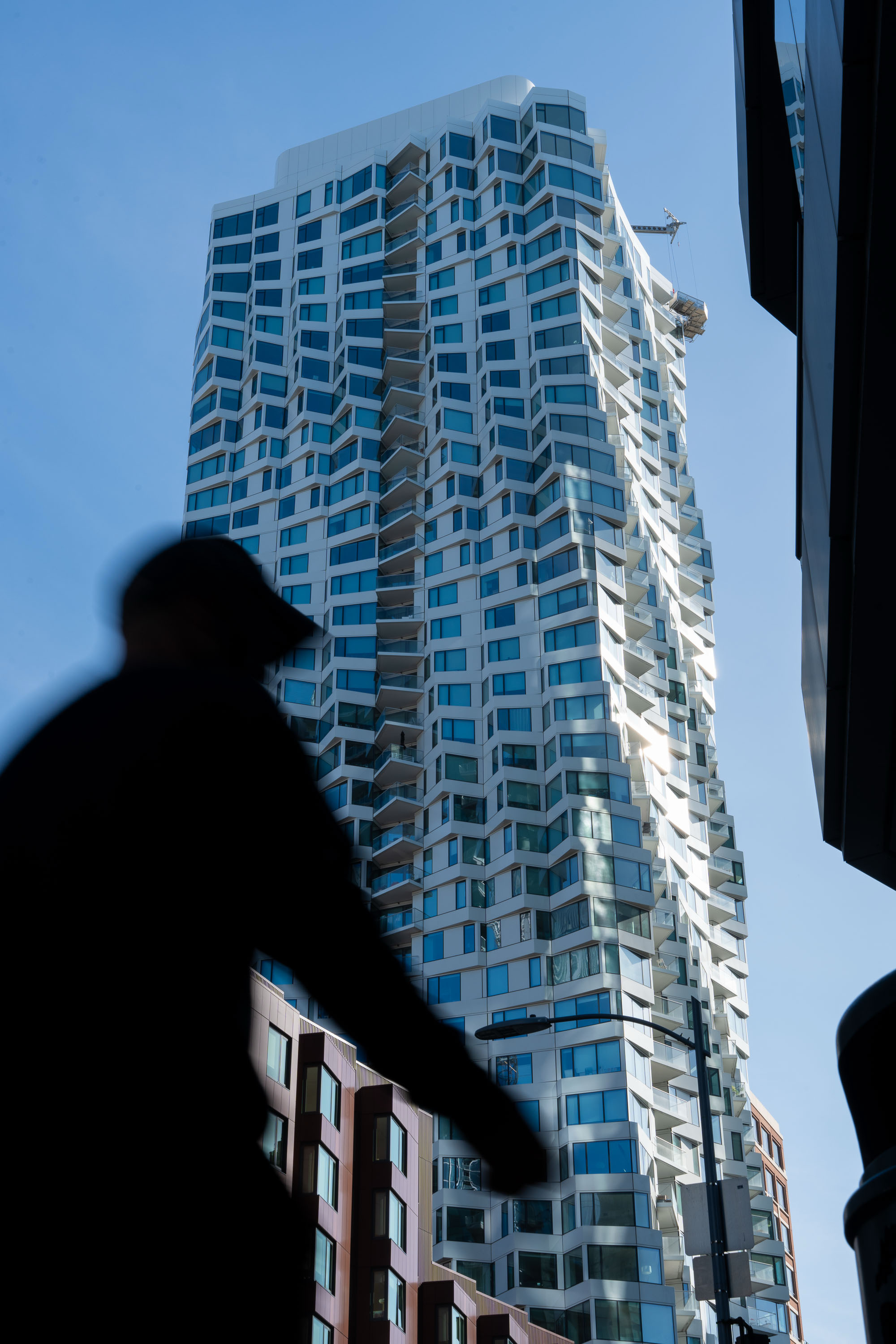 A tall, modern skyscraper with a wavy, geometric design stands against a blue sky. In the foreground, a blurred silhouette of a person is visible.