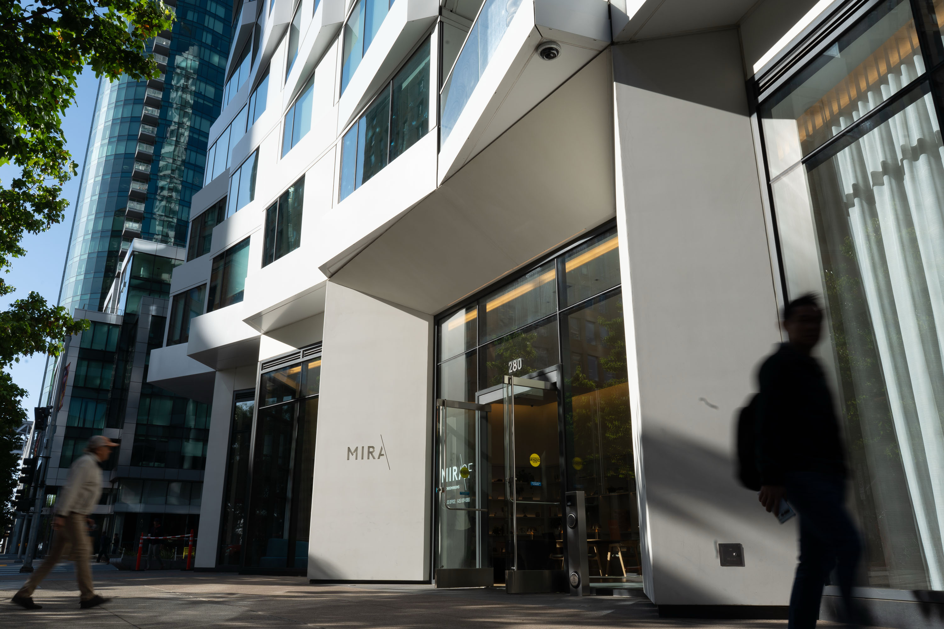 A modern building with a white exterior and large windows, labeled &quot;MIRA,&quot; stands on a sunny street. Two people are walking by, one blurred in motion.