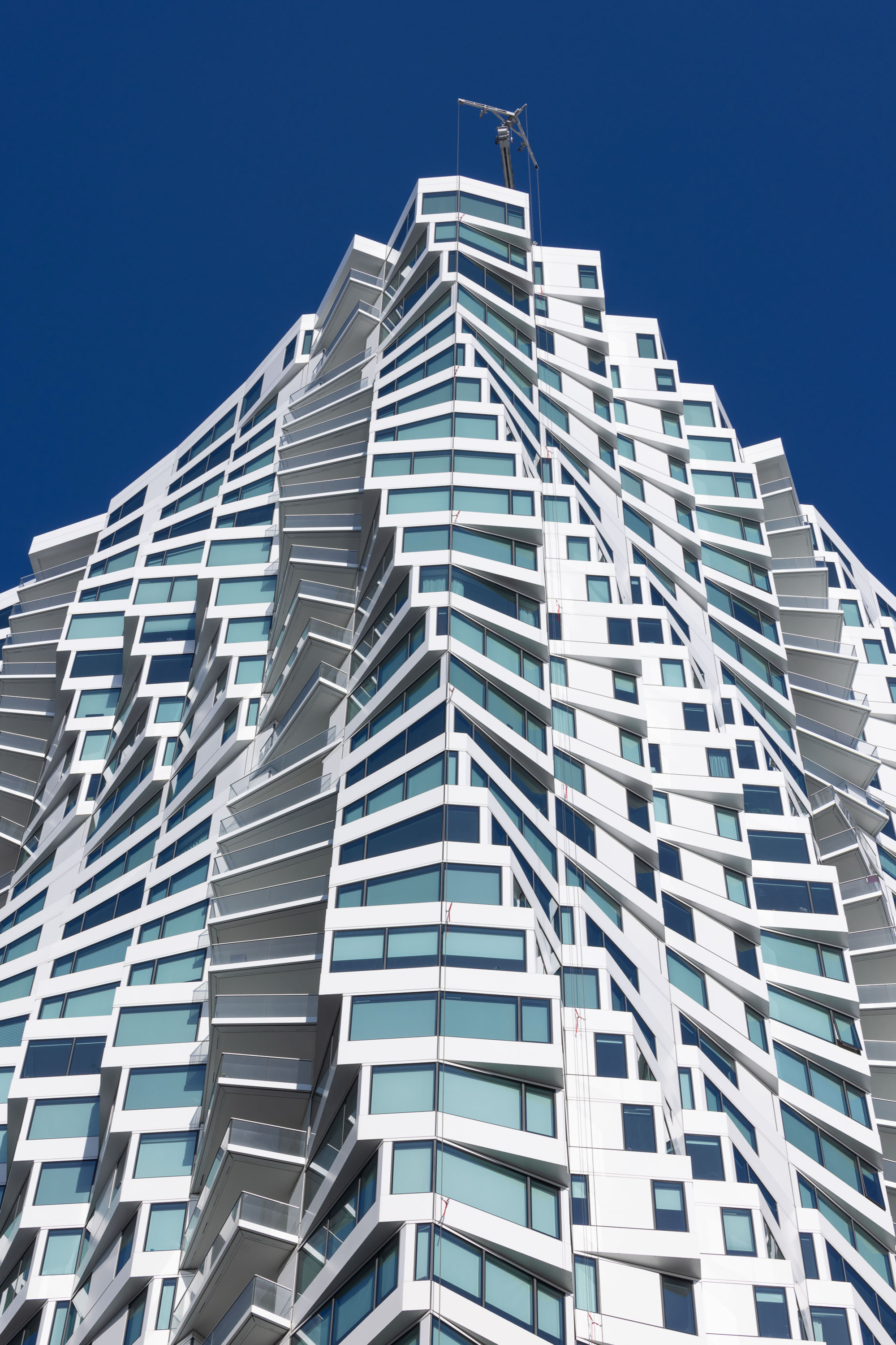 The image shows a modern skyscraper with irregular, zigzagging white balconies and blue-tinted windows against a clear blue sky.