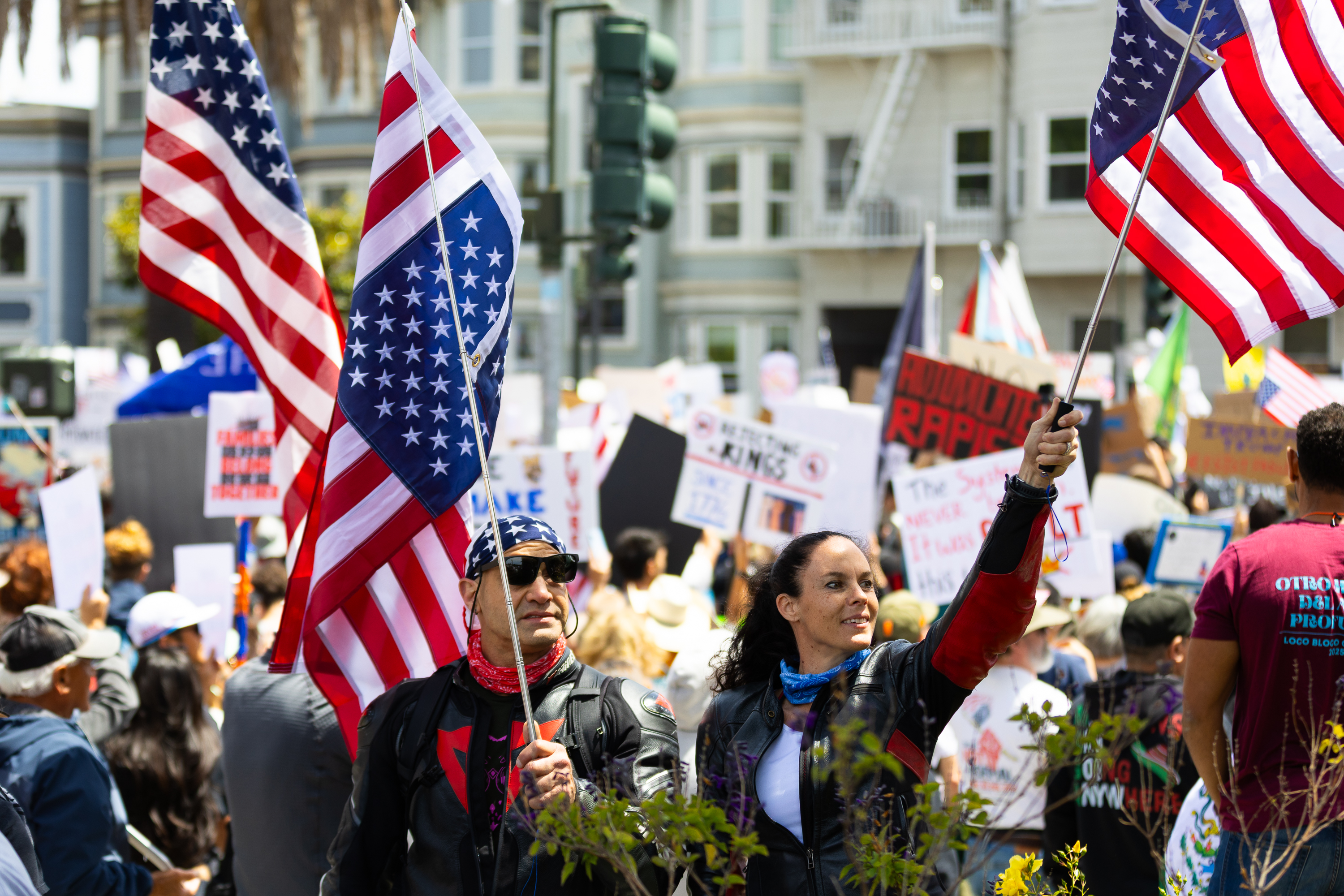 A group of people is participating in a protest or rally, holding numerous American flags and signs. The atmosphere appears lively and busy.