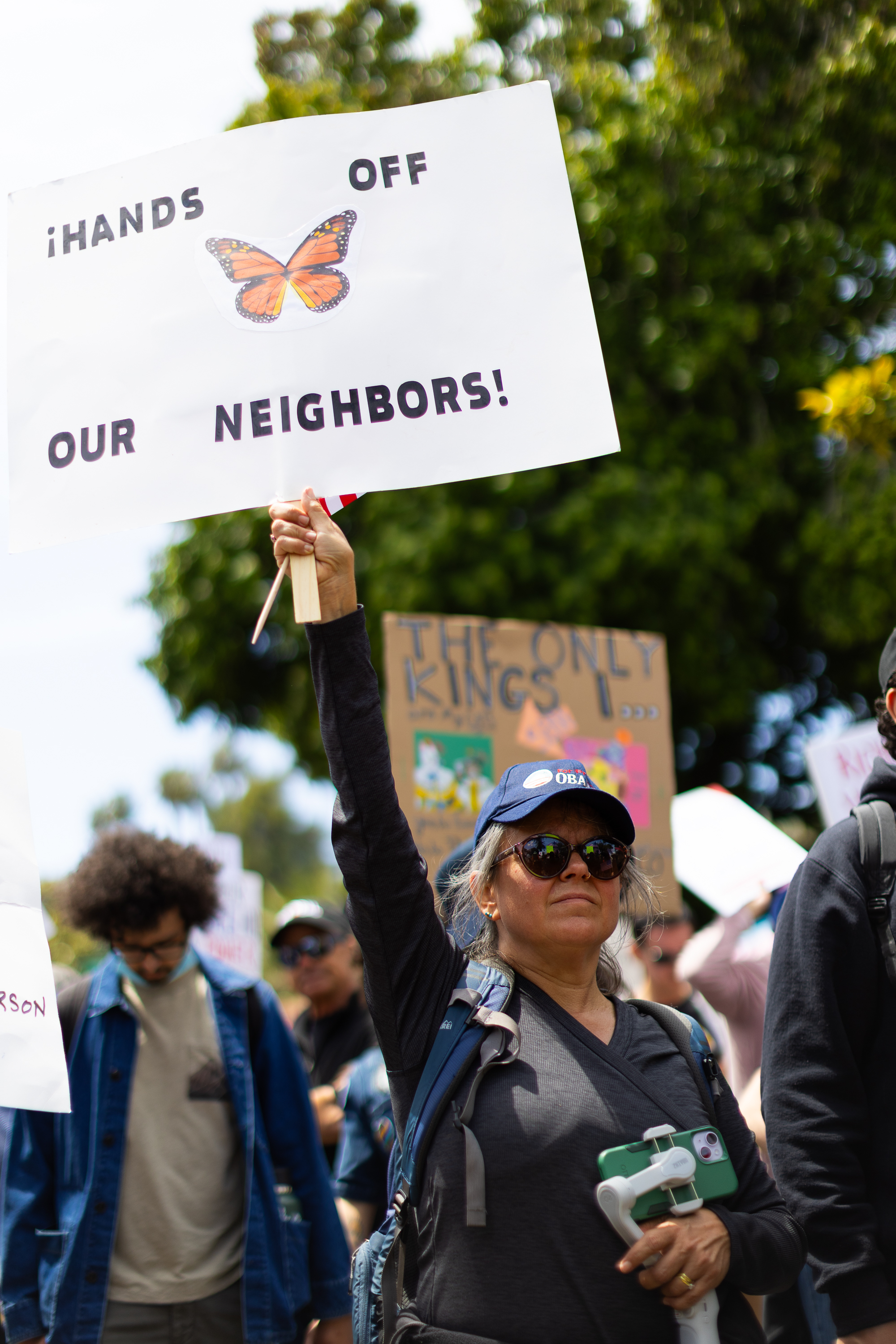 A woman in sunglasses and a cap holds a sign that reads &quot;¡Hands off our neighbors!&quot; with a butterfly image. People are in the background at a rally.