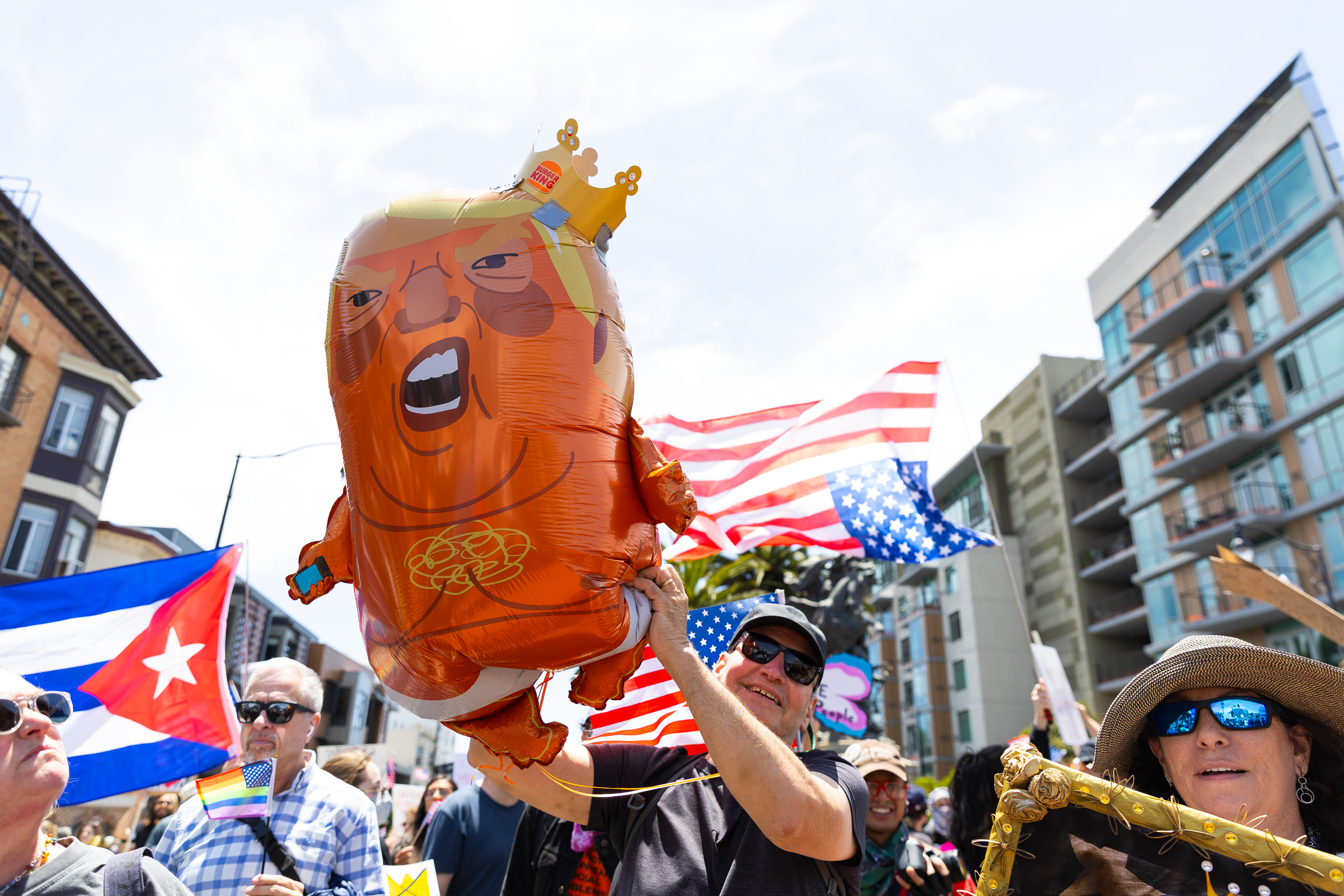 The image shows a crowd at a protest holding a giant balloon caricature and waving flags. People appear spirited, with buildings in the background.