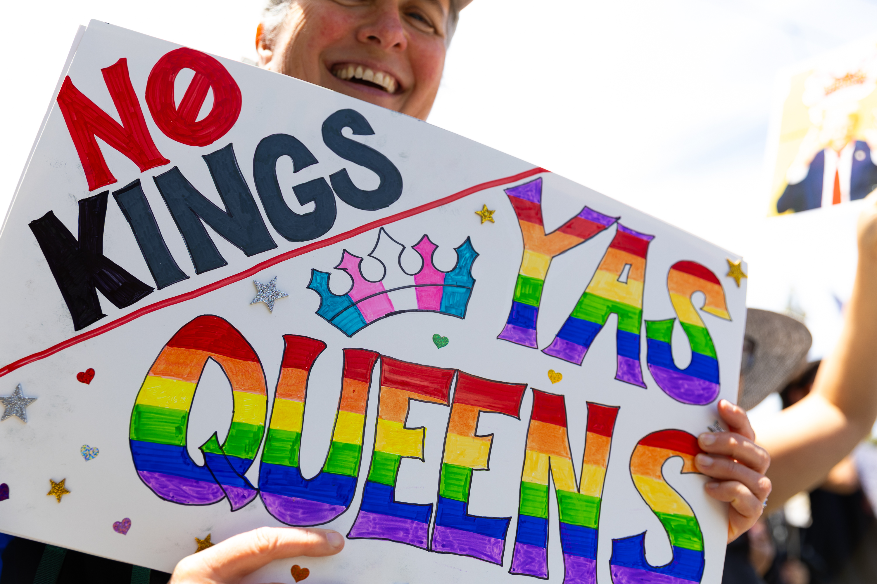 A person is holding a colorful sign reading &quot;No Kings, Yas Queens&quot; in rainbow colors with a crown and stars, expressing pride and empowerment.