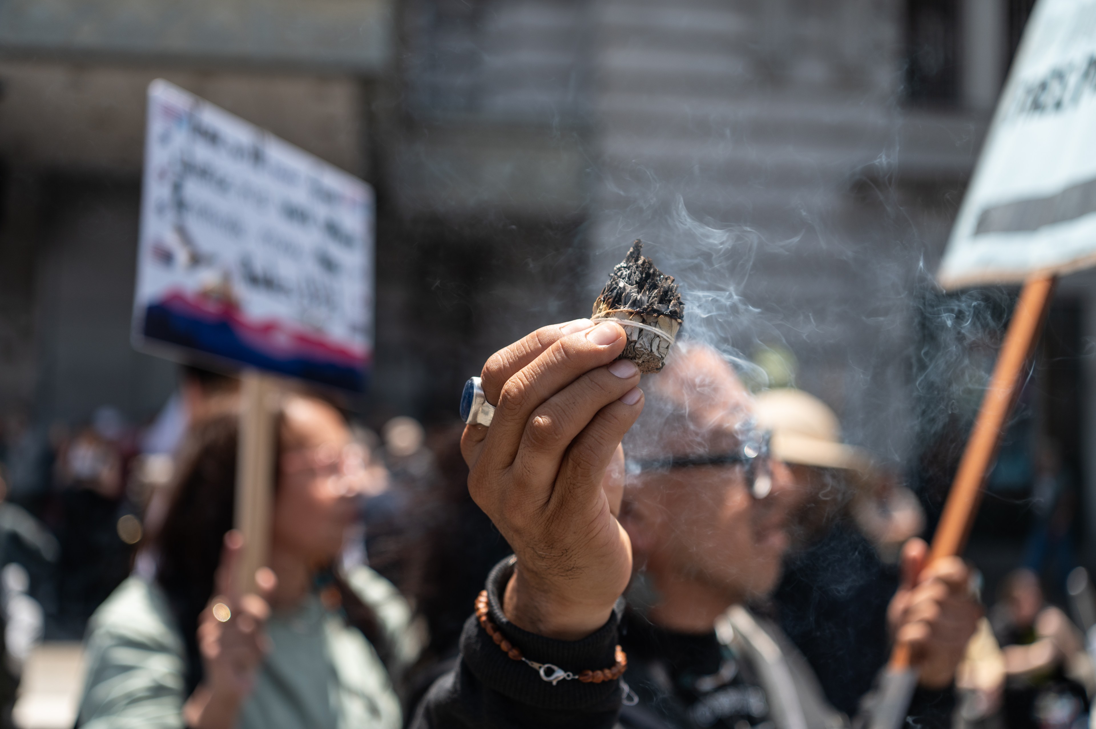 A hand holding a smoking bundle of sage is raised in the foreground. In the background, blurred people hold protest signs. The atmosphere is bustling.