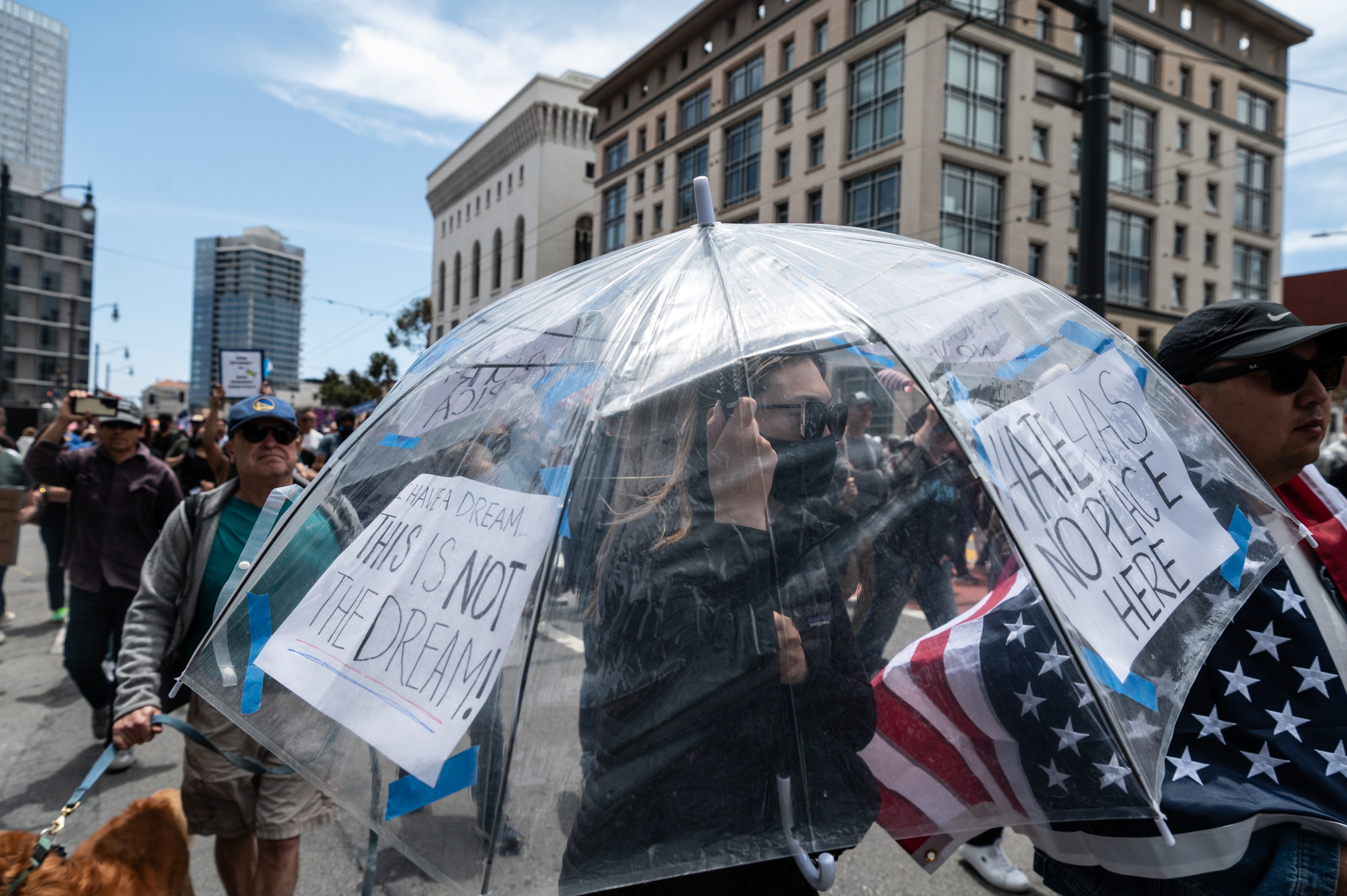 People are marching in a protest. A person holds a transparent umbrella featuring messages like &quot;I have a dream… this is not the dream!&quot; and &quot;Hate has no place here.&quot;
