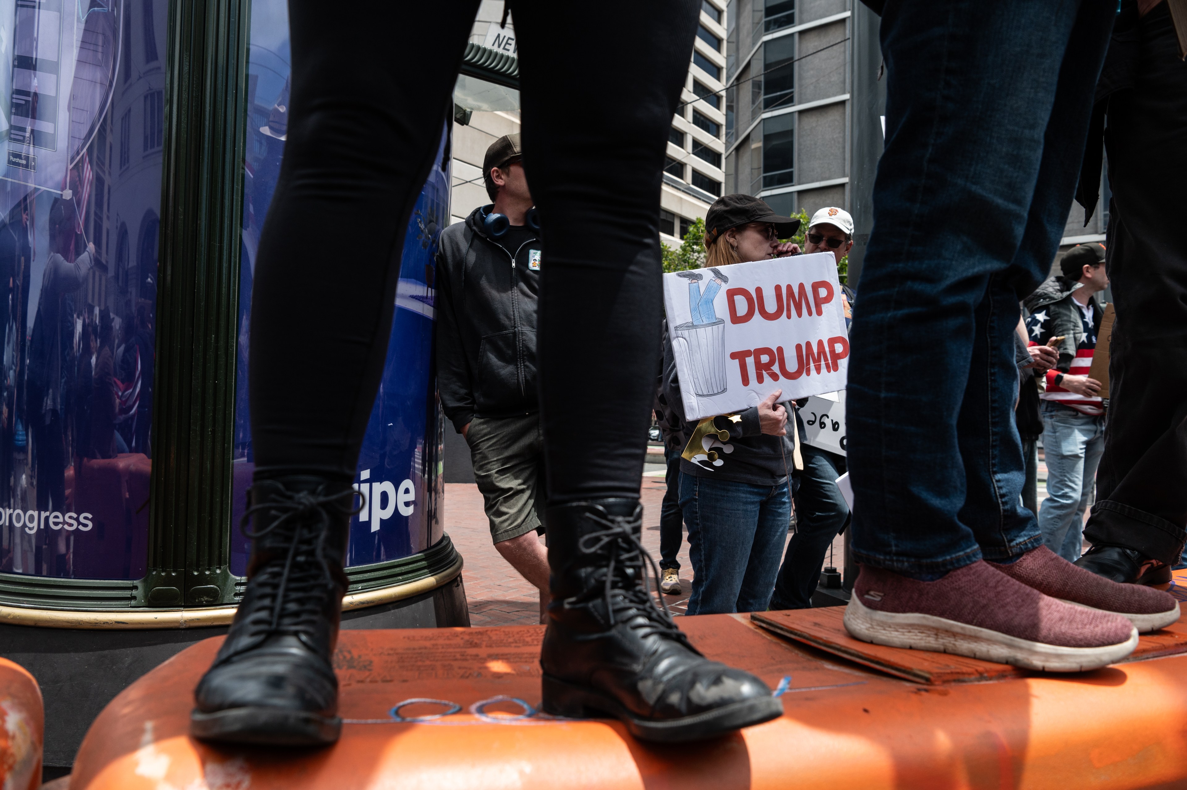 People stand on an orange barrier at a protest. One holds a sign reading &quot;Dump Trump,&quot; depicting a figure being tossed into a trash can.