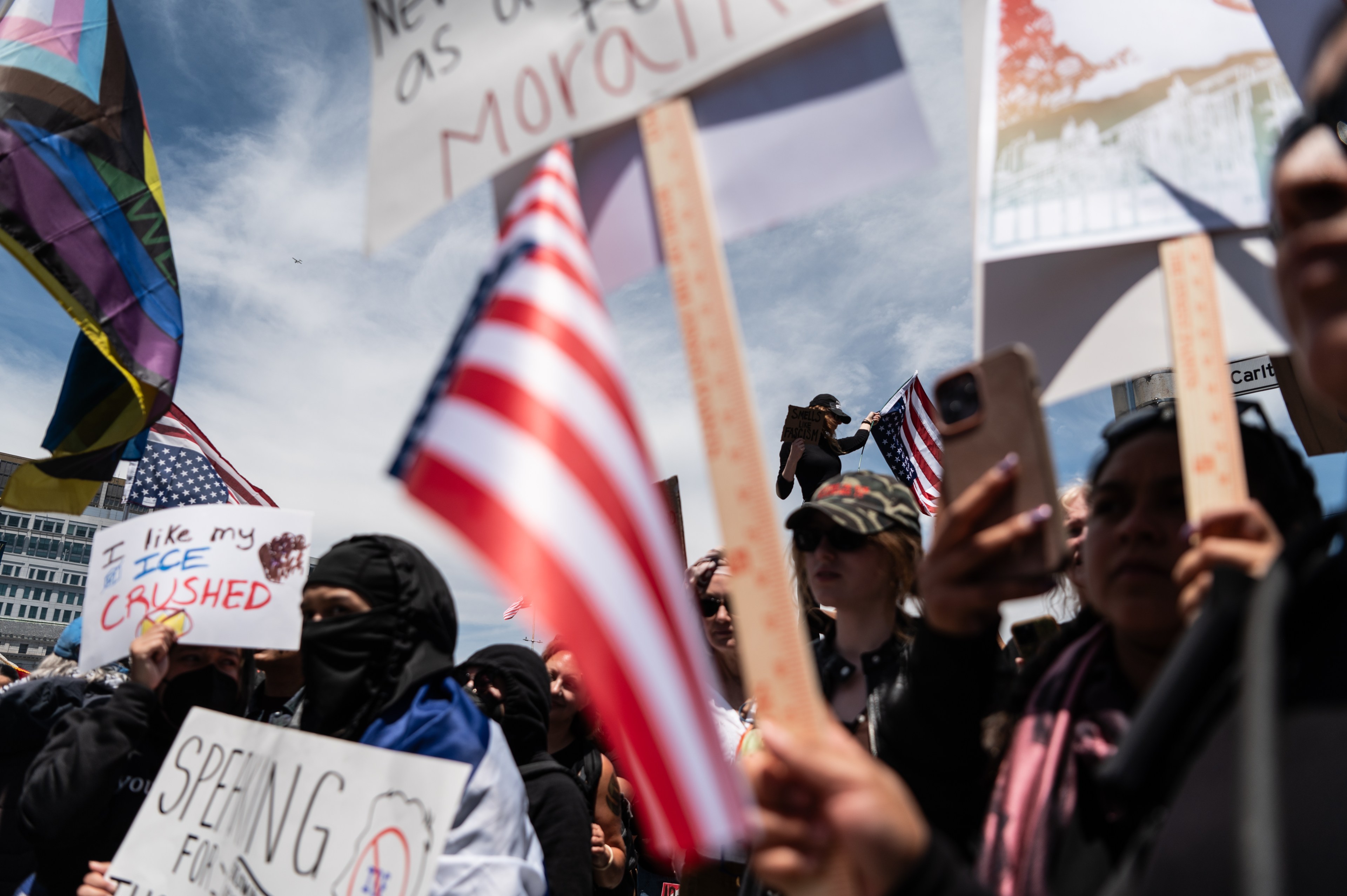 The image shows a lively protest with people holding signs and American flags. The crowd is diverse, and the atmosphere seems energetic and determined.
