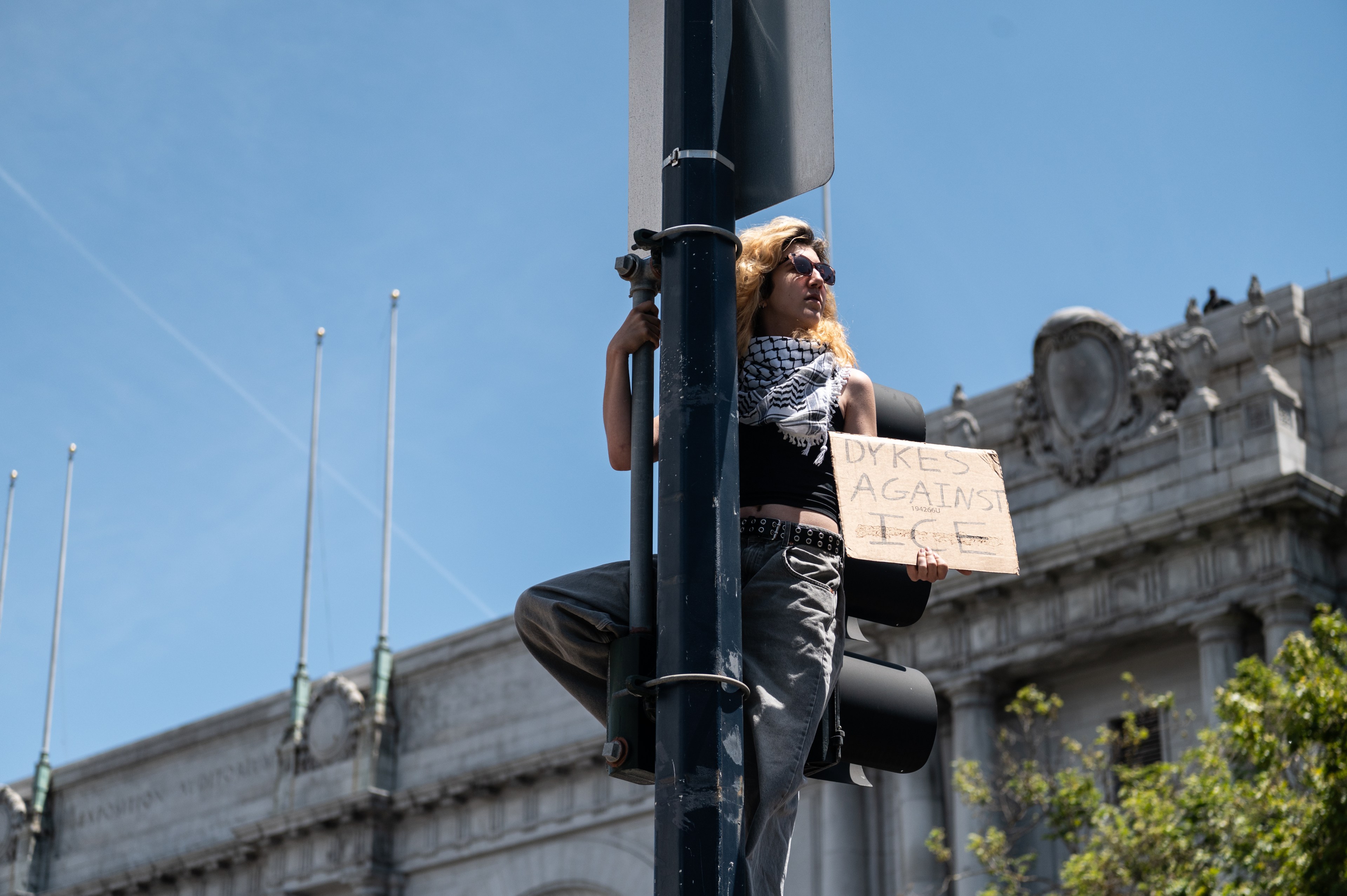 A person with long hair is climbing a pole, holding a cardboard sign reading &quot;Dykes Against ICE.&quot; They are wearing sunglasses and a black and white scarf.