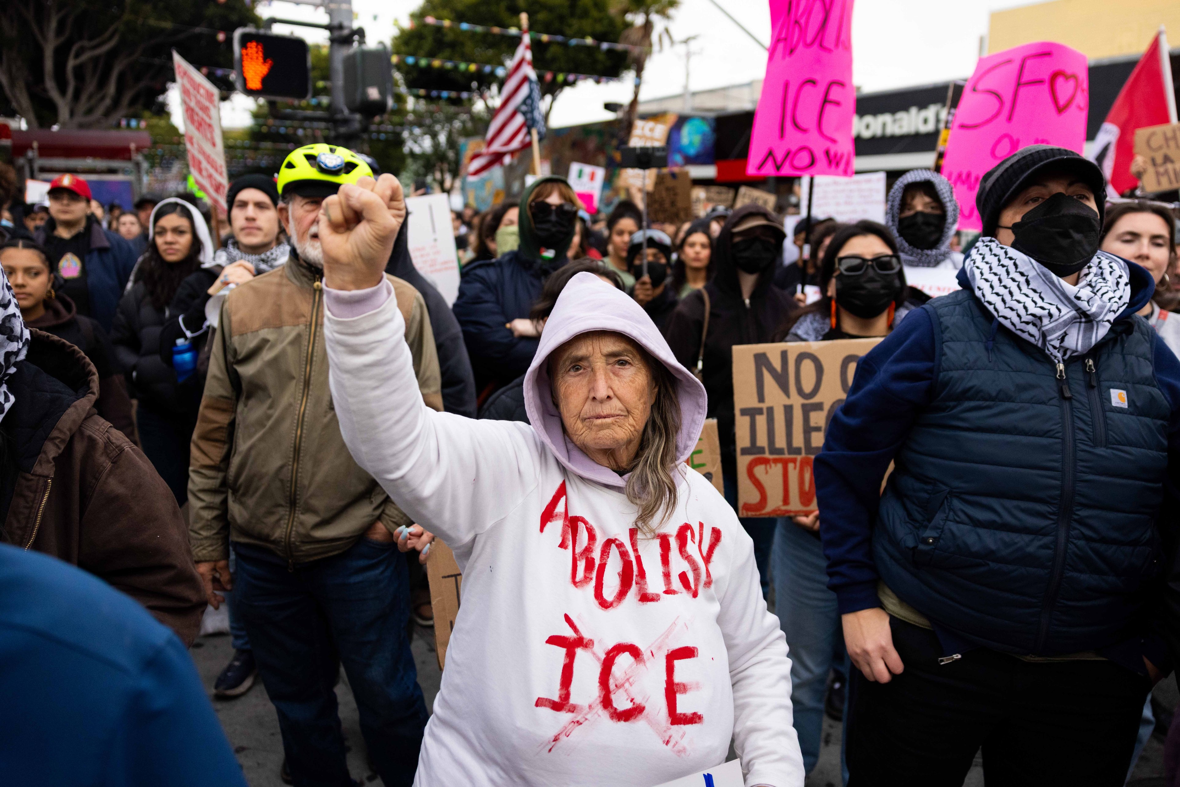 A crowd of protesters is marching, many holding signs. An older woman in a white hoodie with "Abolish ICE" written on it raises her fist in the foreground.