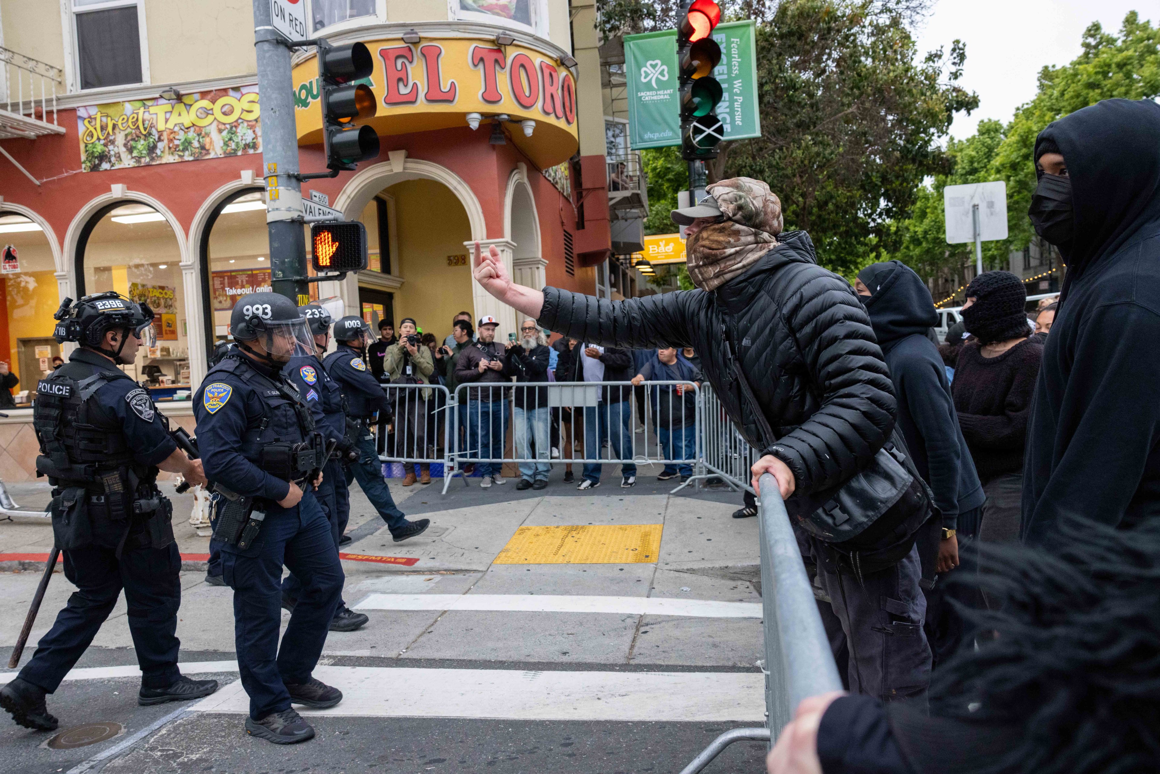 A person gestures towards police officers in riot gear standing near metal barricades on a street corner. A crowd observes from behind the barricades.