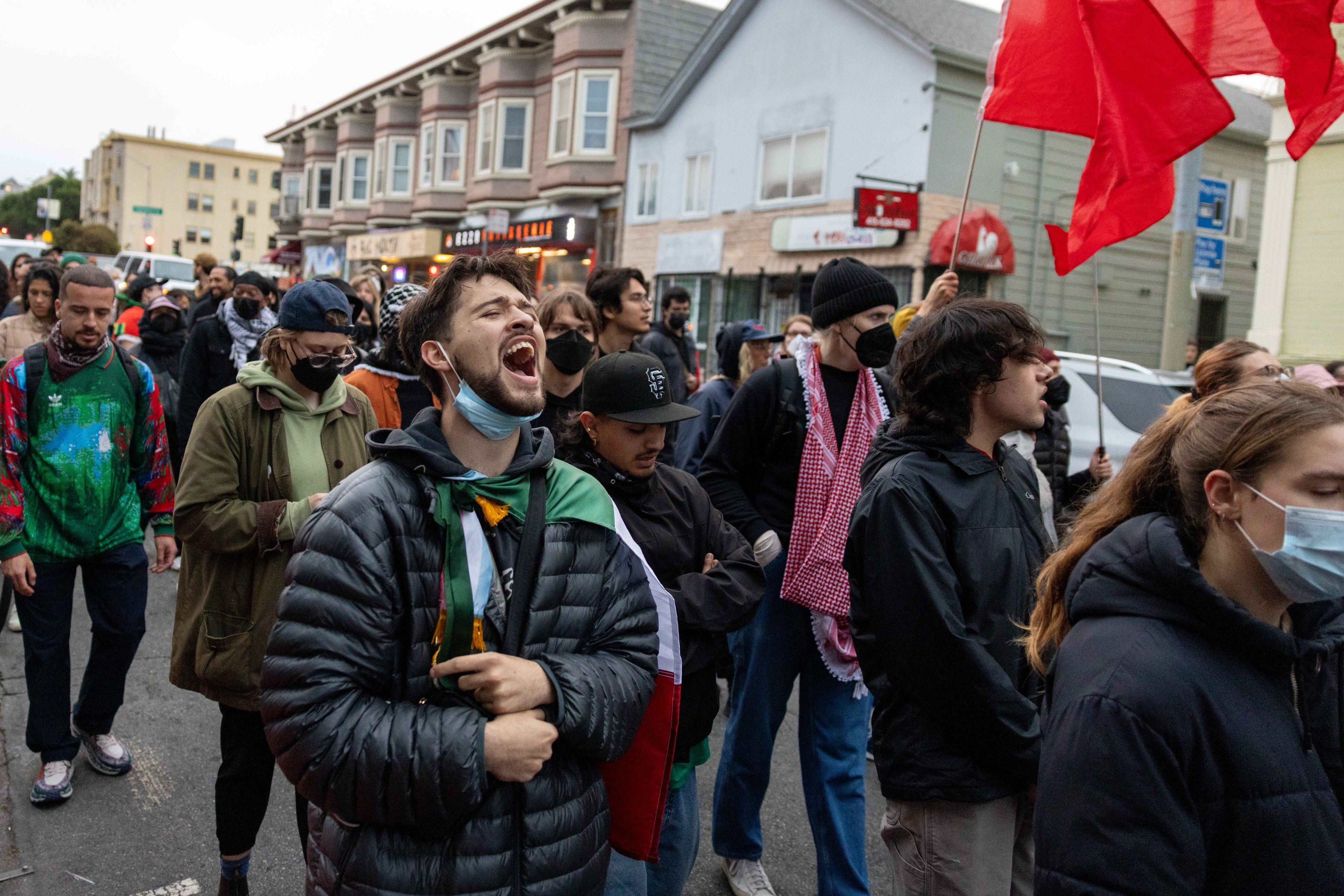 A crowd of people is participating in a street protest. Some wear masks and hold red flags, and a person in the foreground appears to be shouting.