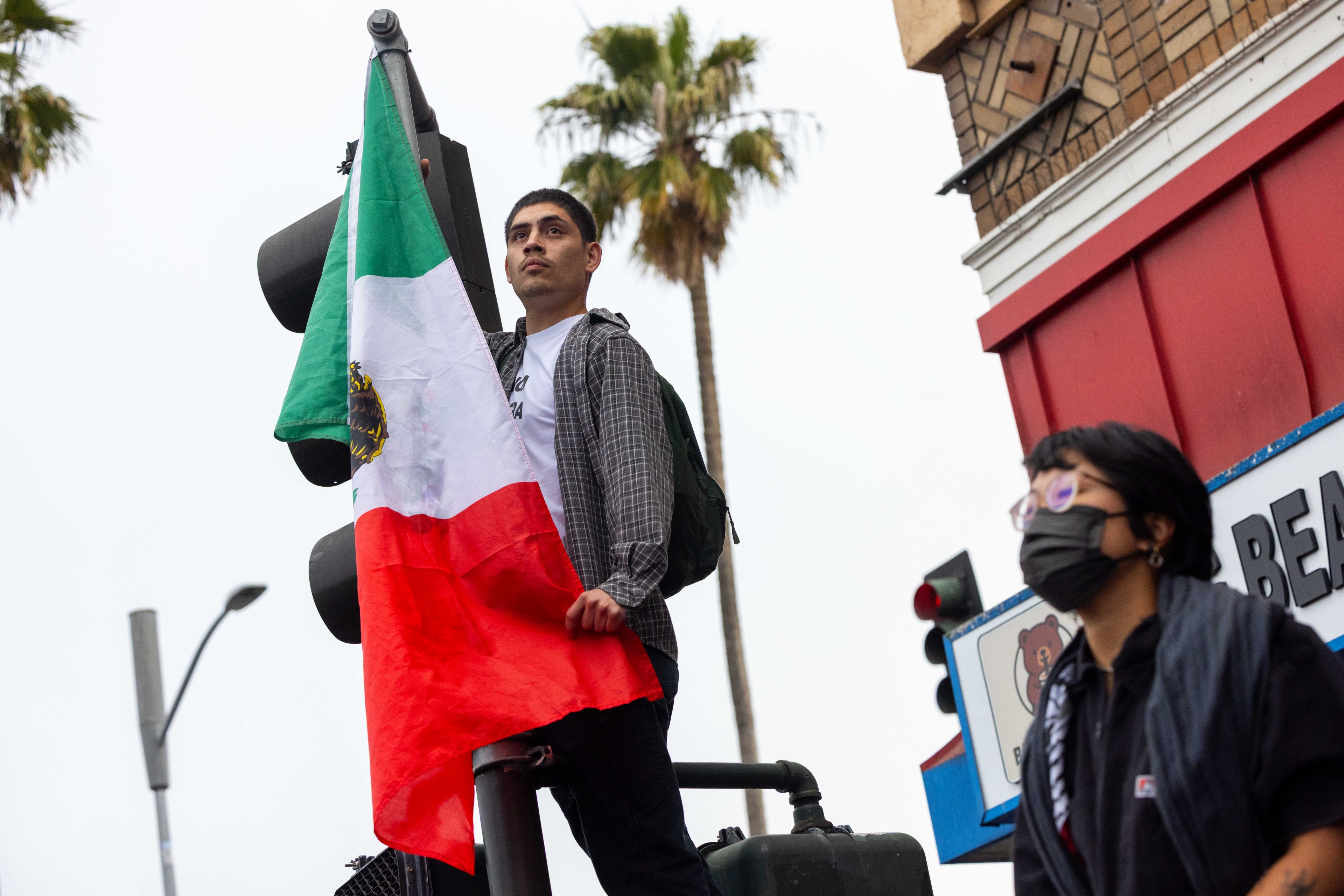 A person stands on a traffic light holding a Mexican flag. Another person in a mask walks nearby. Palm trees and a building are in the background.