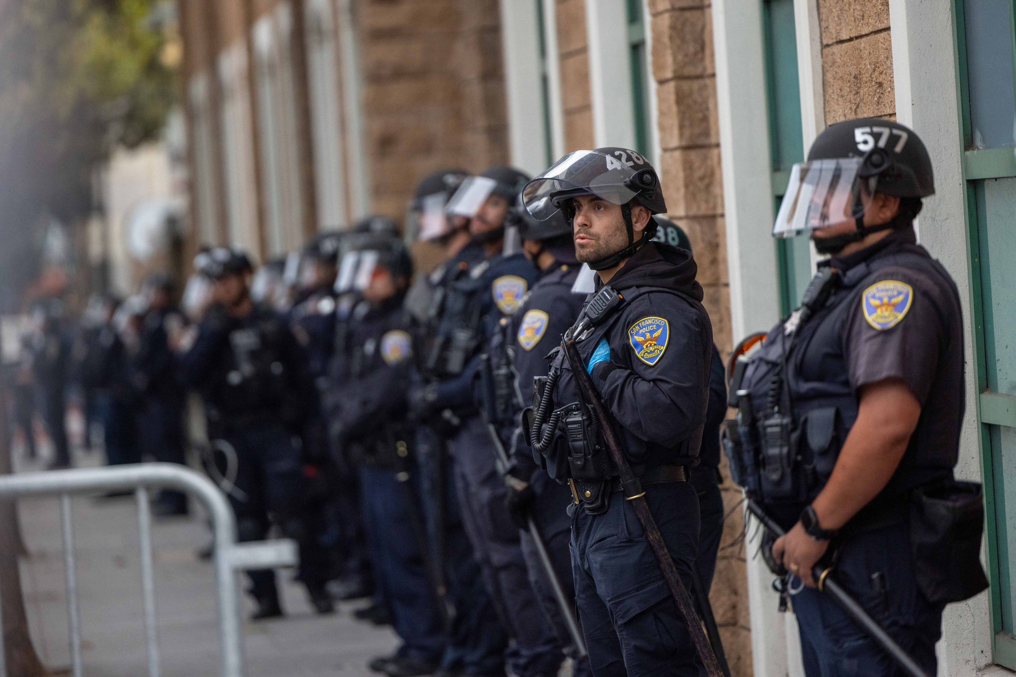 A line of police officers in riot gear, wearing helmets and face shields, stands against a building. They appear alert, holding batons, with focus on one officer.