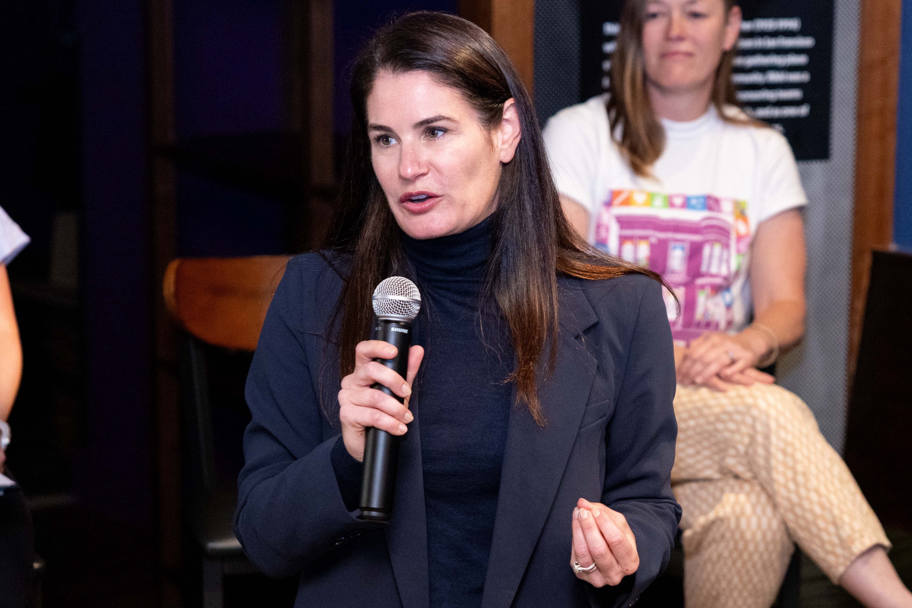 A woman with long brown hair holds a microphone, speaking intently. She's wearing a dark jacket. Another woman sits in the background, watching.