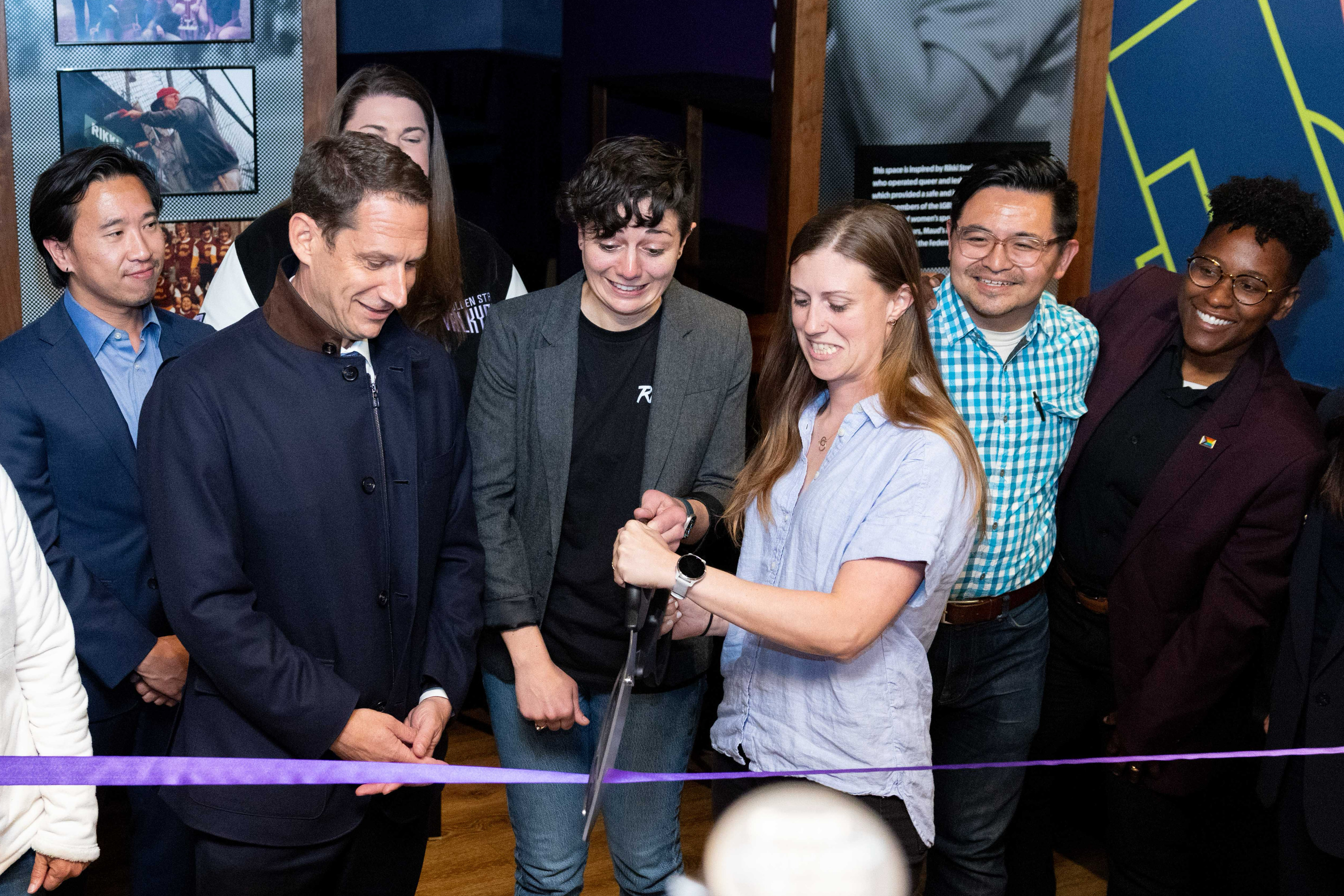 A group of people gather as a woman cuts a purple ribbon with large scissors, marking a ceremonial event. They all appear cheerful and engaged.