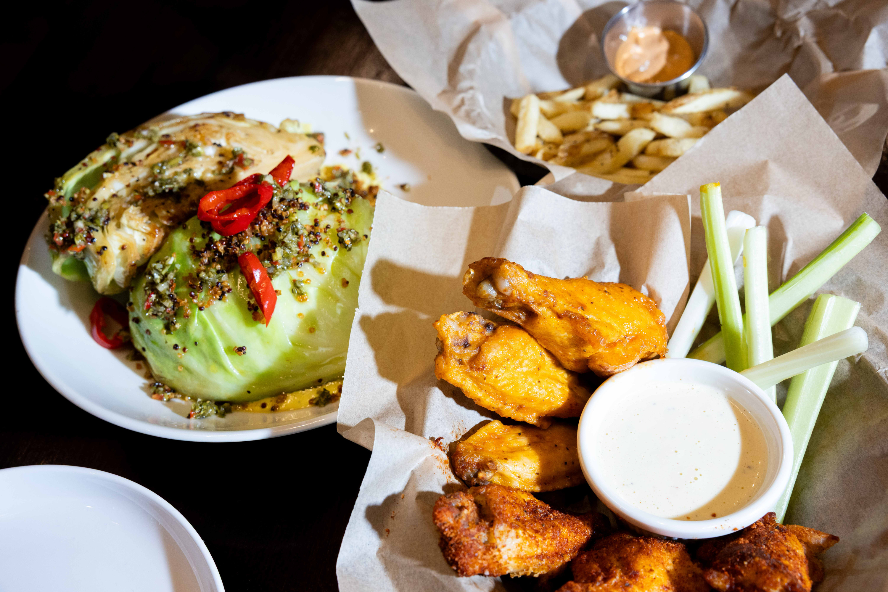 A plate of lettuce wedges with red peppers, a basket of chicken wings with a ranch dip and celery sticks, and fries with sauce in the background.