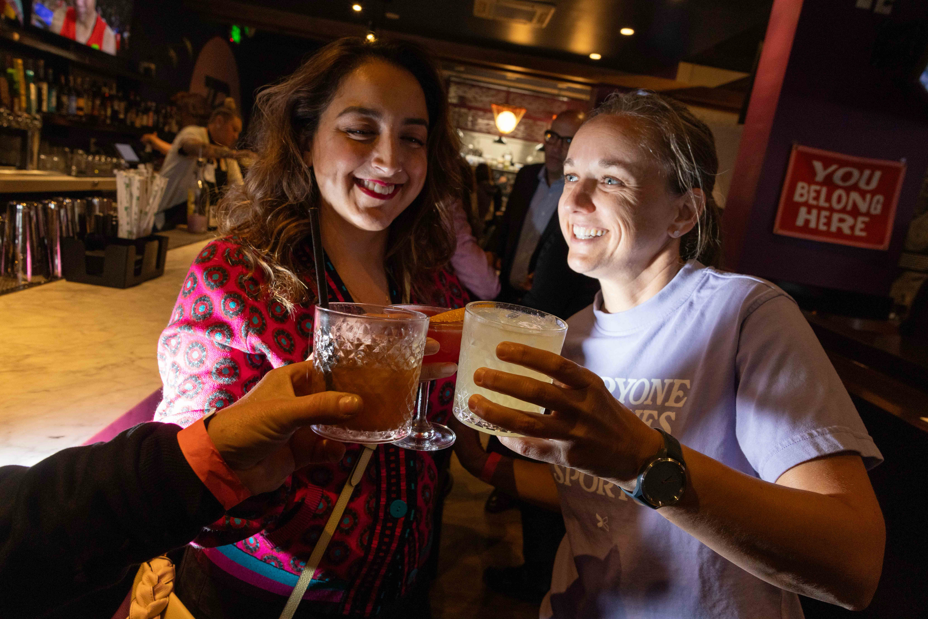 Two women are smiling and clinking glasses with someone at a bar, where a sign reads &quot;You Belong Here.&quot; The atmosphere is lively and welcoming.