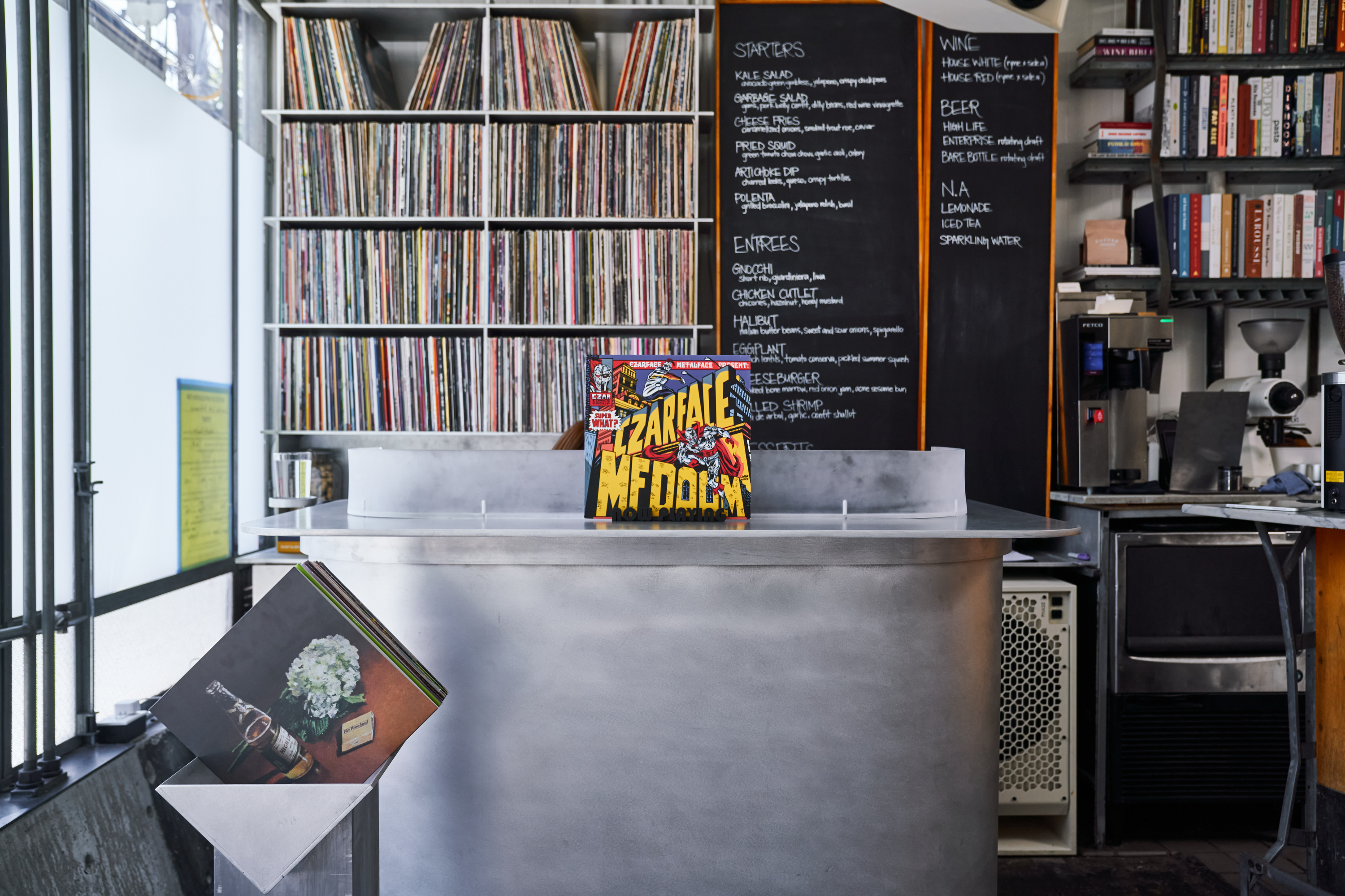 A small, modern cafe interior with a metal counter, menu board on the wall, shelves of records, books, coffee maker, and vibrant album covers displayed.