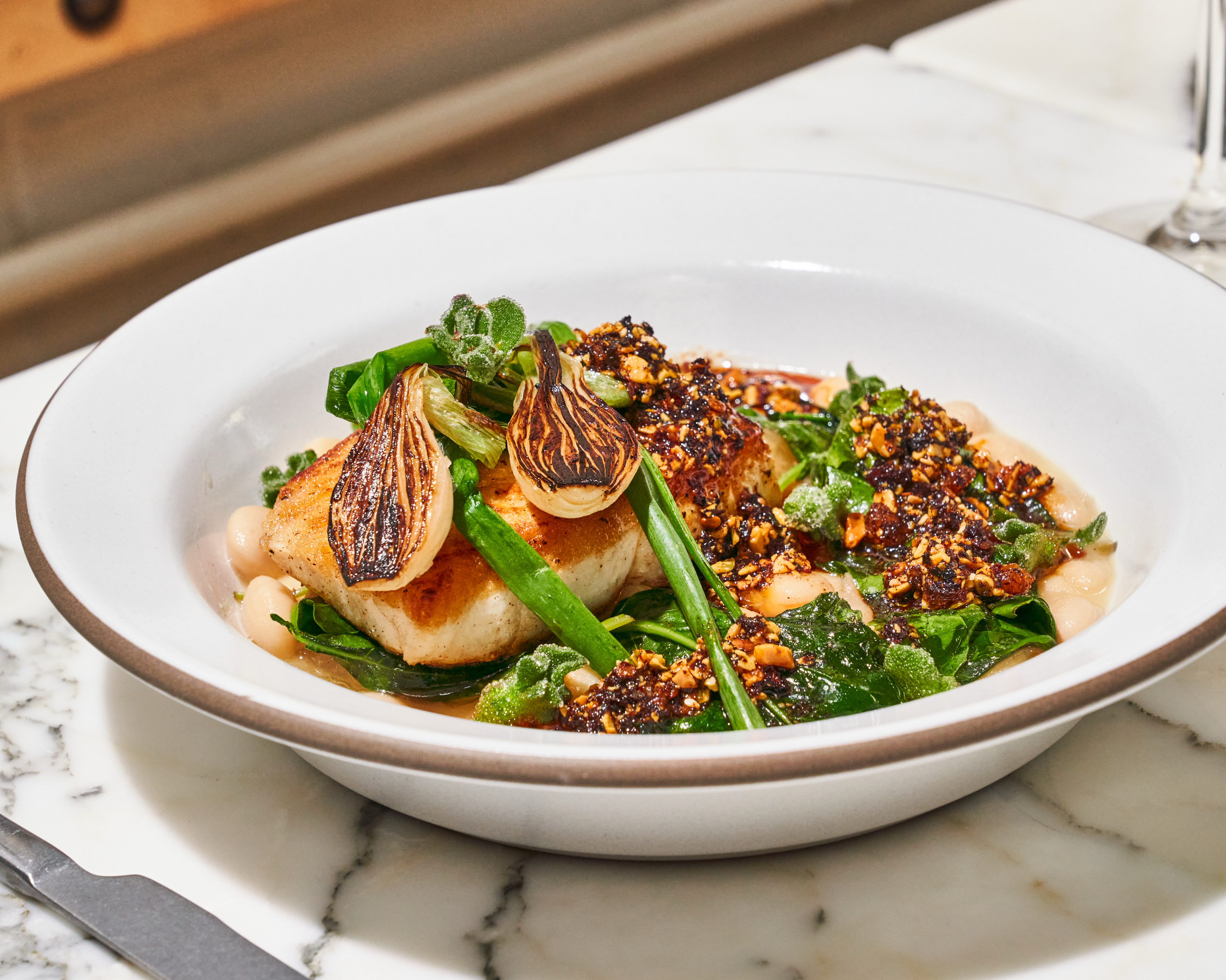 A white plate contains seared fish topped with roasted onions, nuts, and greens, all resting on a bed of creamy white beans on a marble surface.