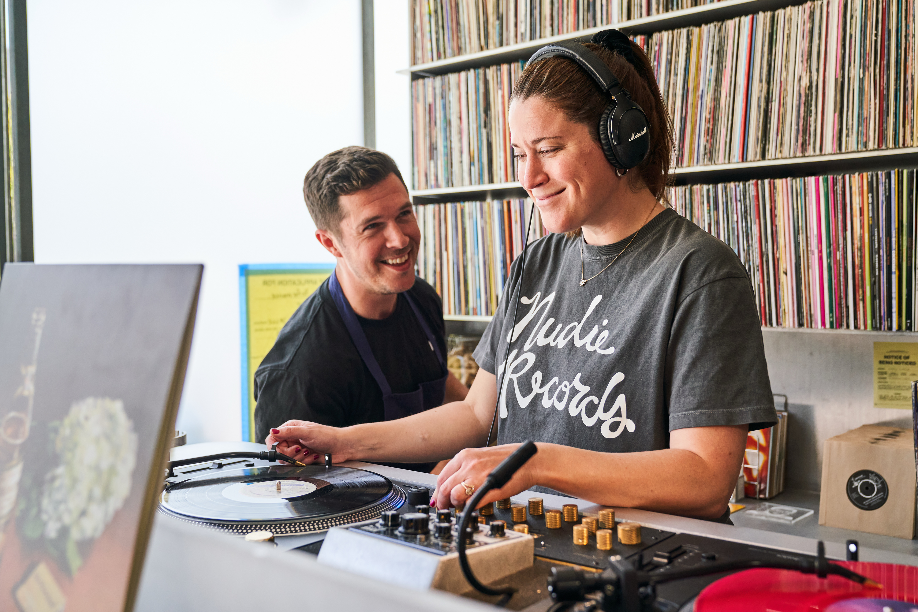 A woman with headphones is DJing, smiling as she adjusts the record. A man beside her looks on happily. Shelves of records are visible in the background.