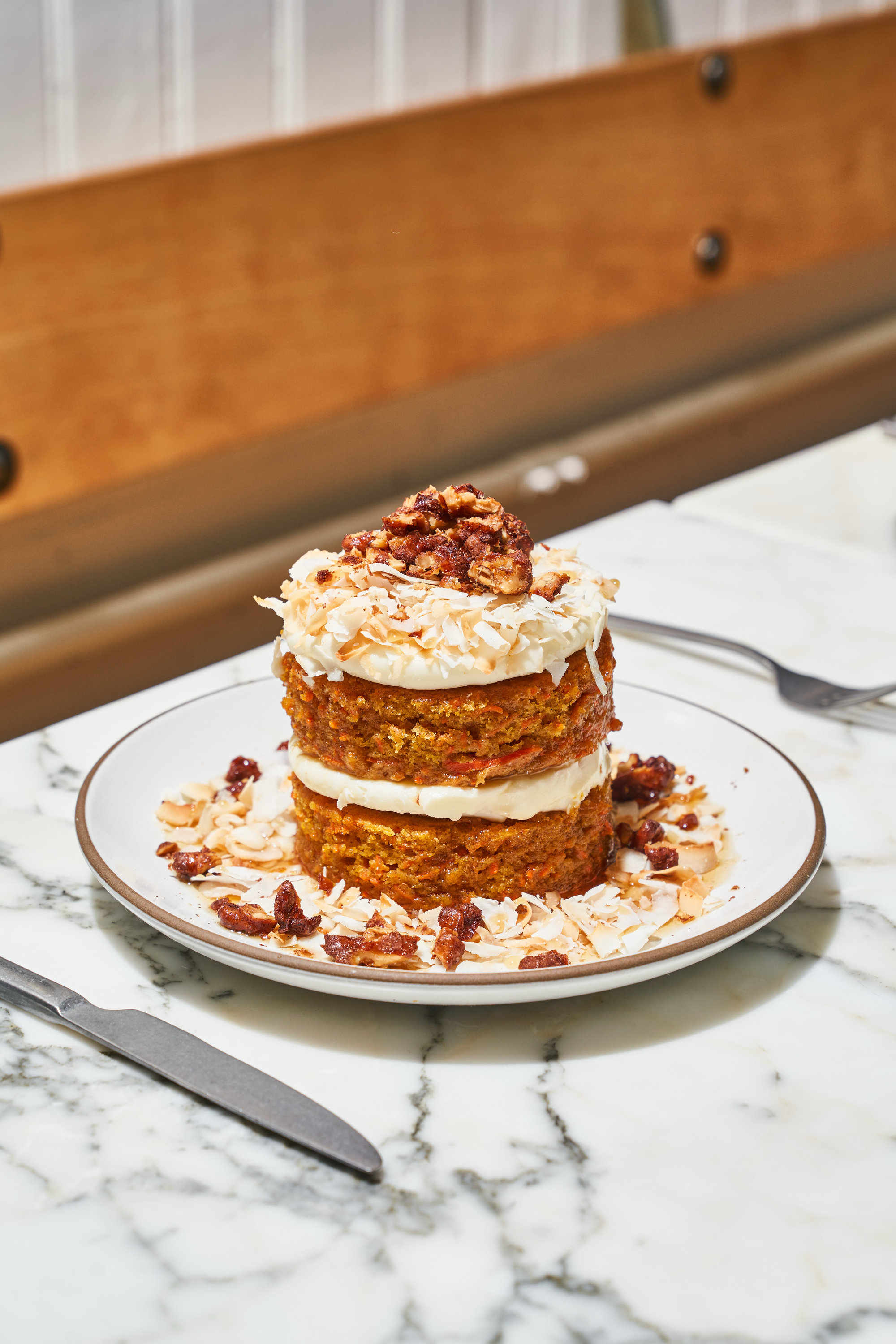 A two-layer carrot cake with cream cheese frosting and nuts sits on a plate, garnished with toasted coconut flakes, on a marble table.