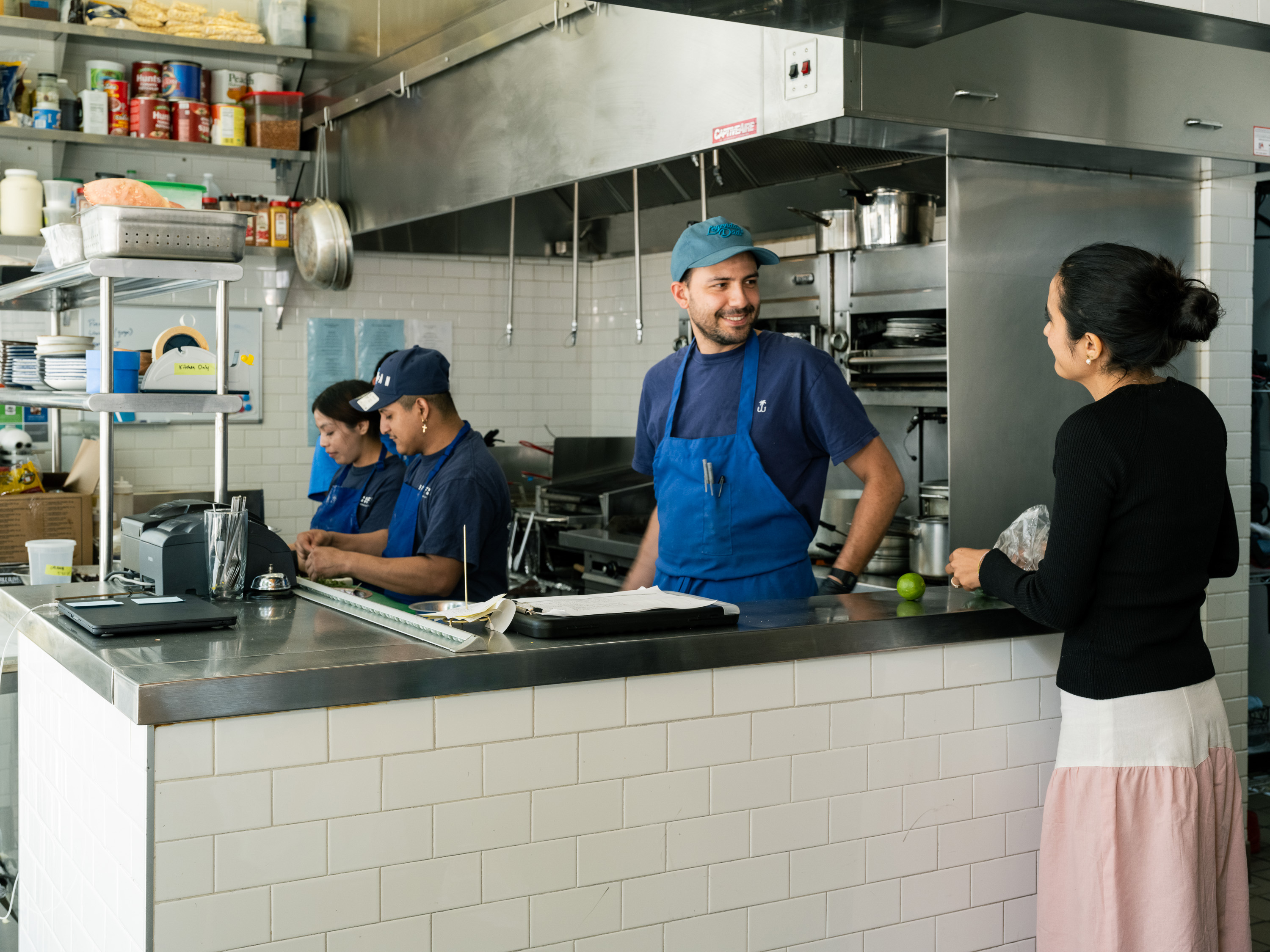 A busy kitchen scene shows three staff members in blue uniforms working, while a woman in a black sweater and skirt smiles, holding a wrapped item at the counter.