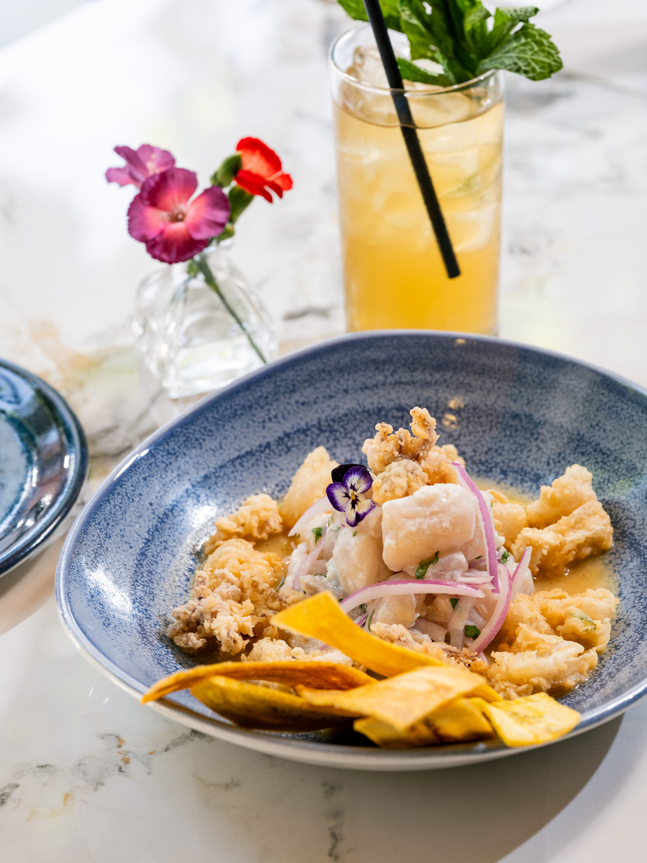A blue bowl with seafood ceviche, garnished with onion, flowers, and plantain chips, sits on a marble table next to a floral vase and a drink with mint leaves.