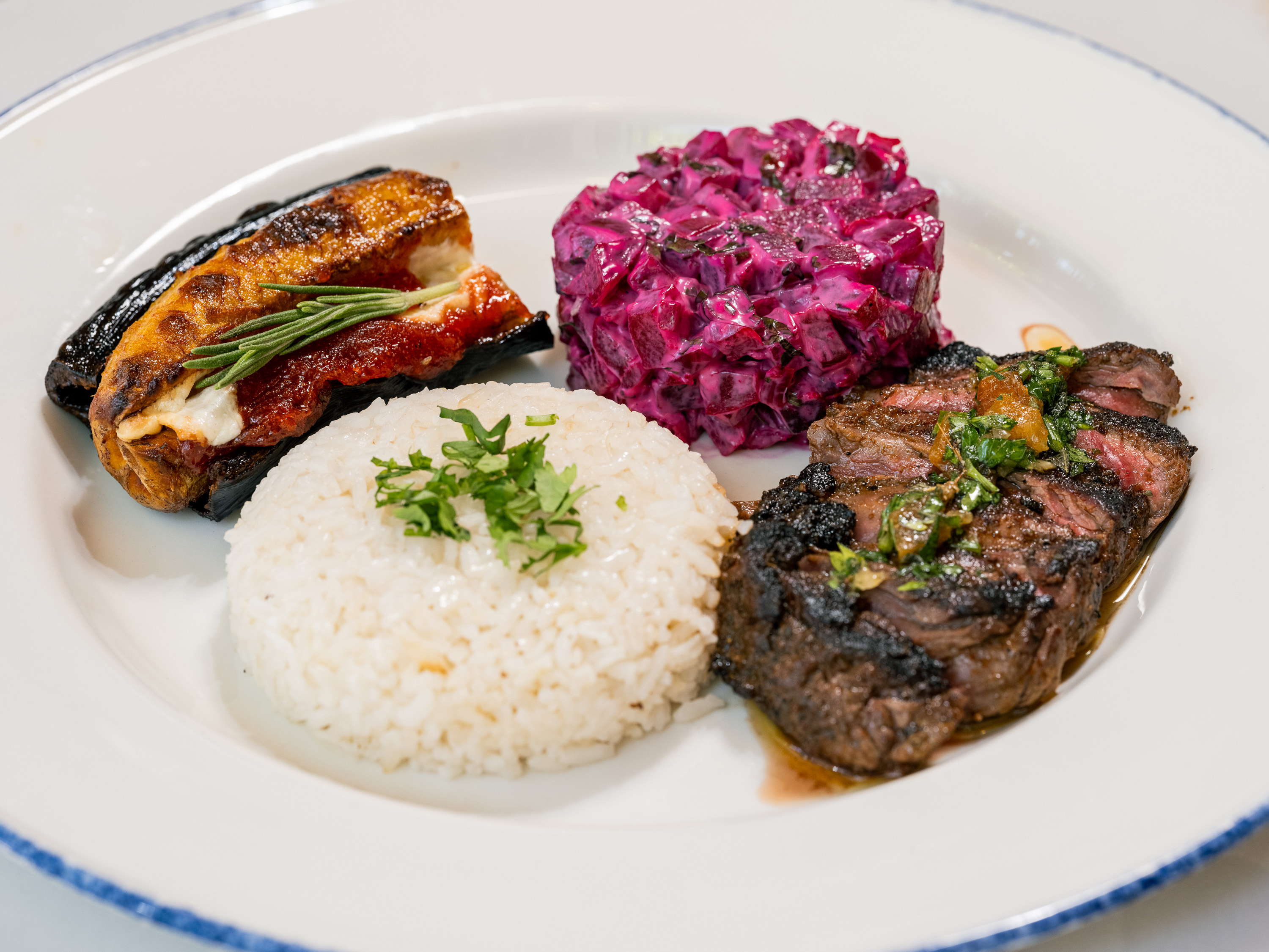 The image shows a plate with grilled meat, a scoop of rice, beetroot salad, and stuffed eggplant, garnished with herbs.