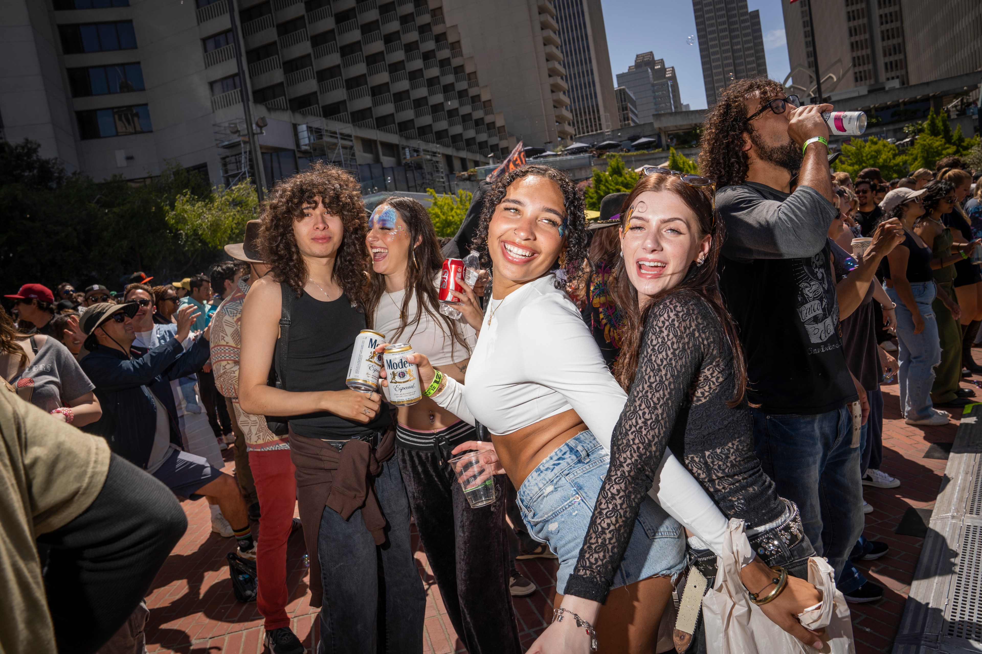 A joyful group is dancing outside at a lively event. They hold drinks and are surrounded by a crowd under clear blue skies, with buildings in the background.