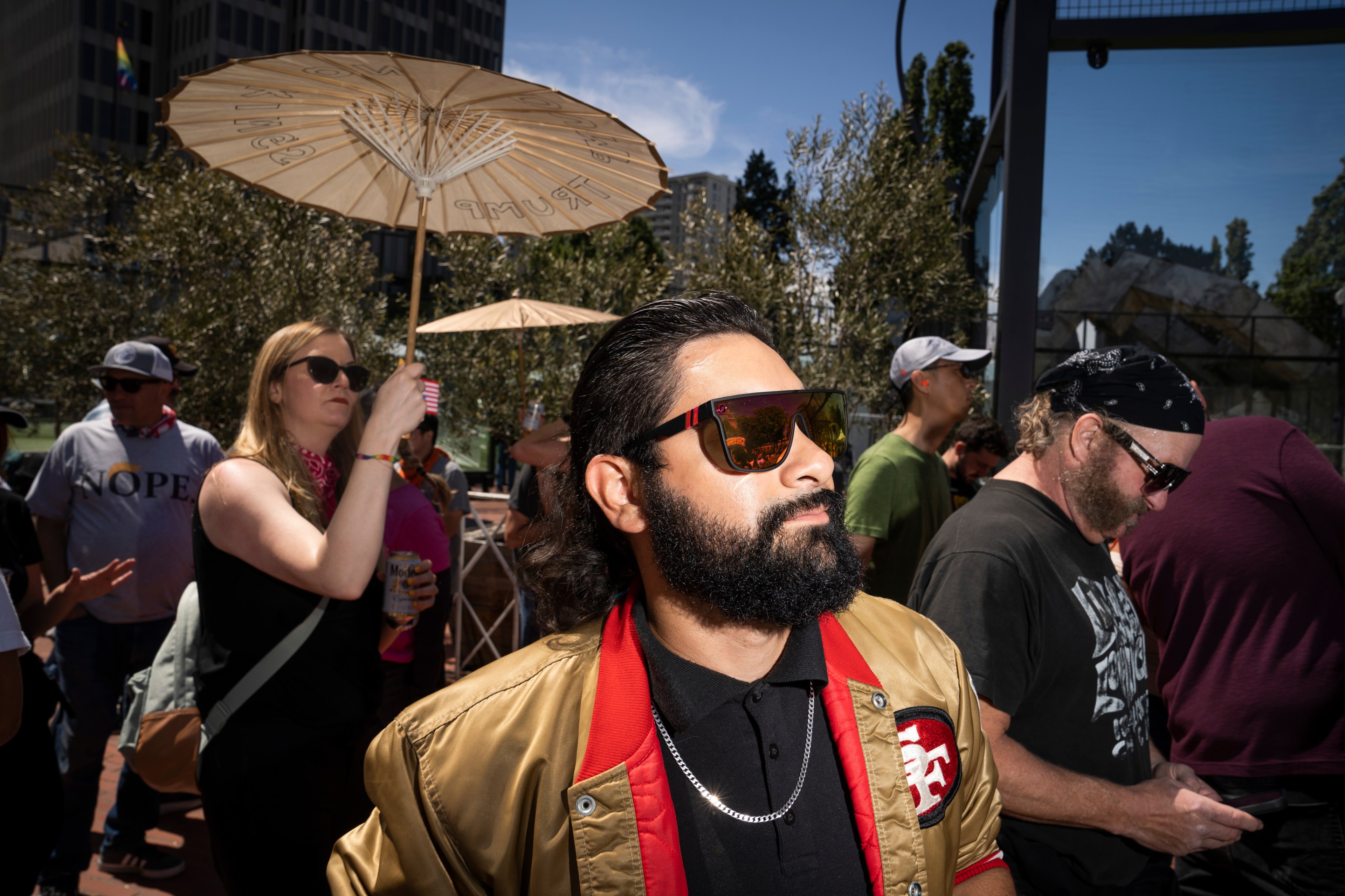 A bearded man in sunglasses and a gold jacket is in the foreground, with people around him holding umbrellas and drinks, under a clear blue sky.