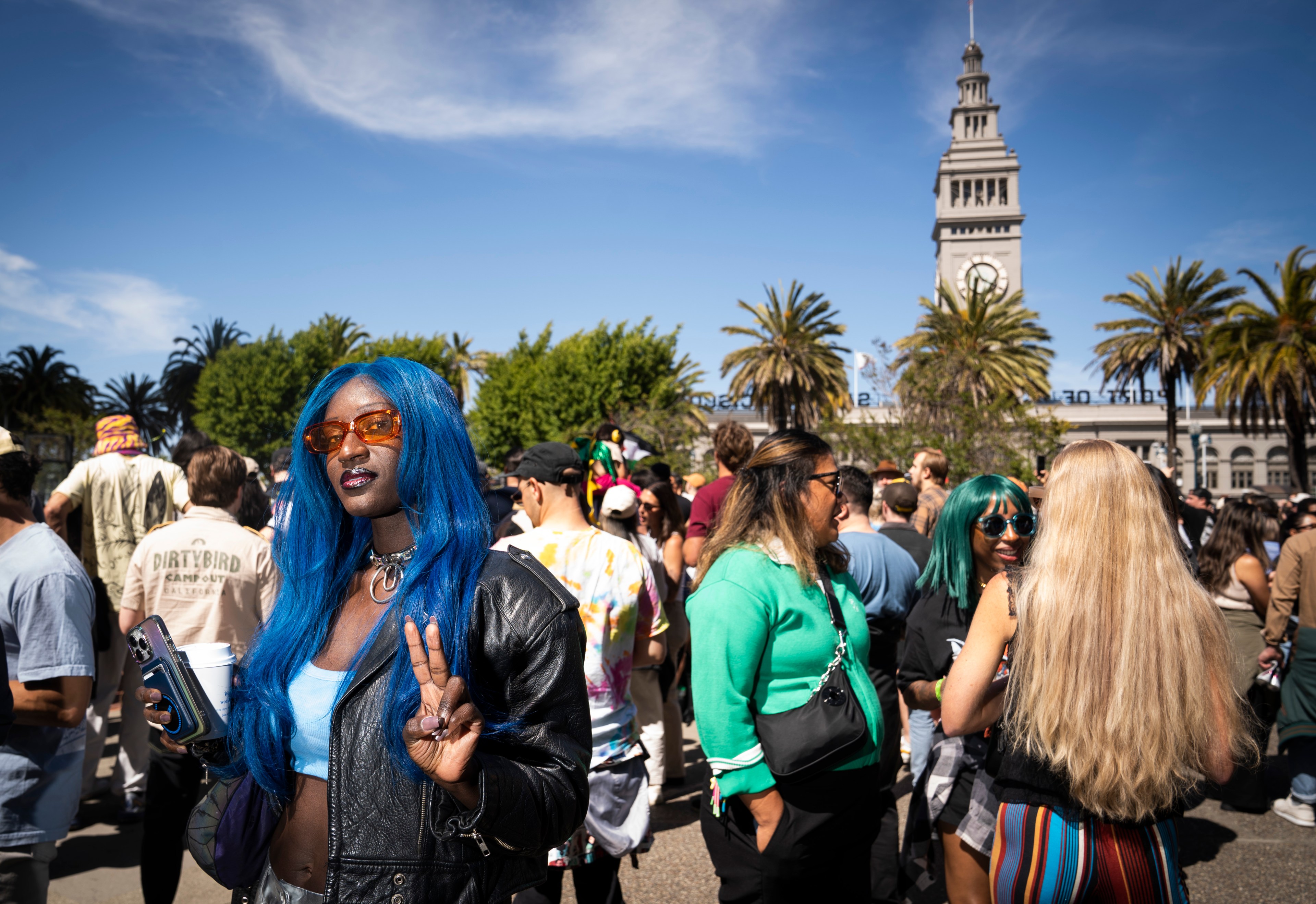 A person with long blue hair and orange sunglasses flashes a peace sign at a crowded outdoor event. A clock tower and palm trees are visible in the background.