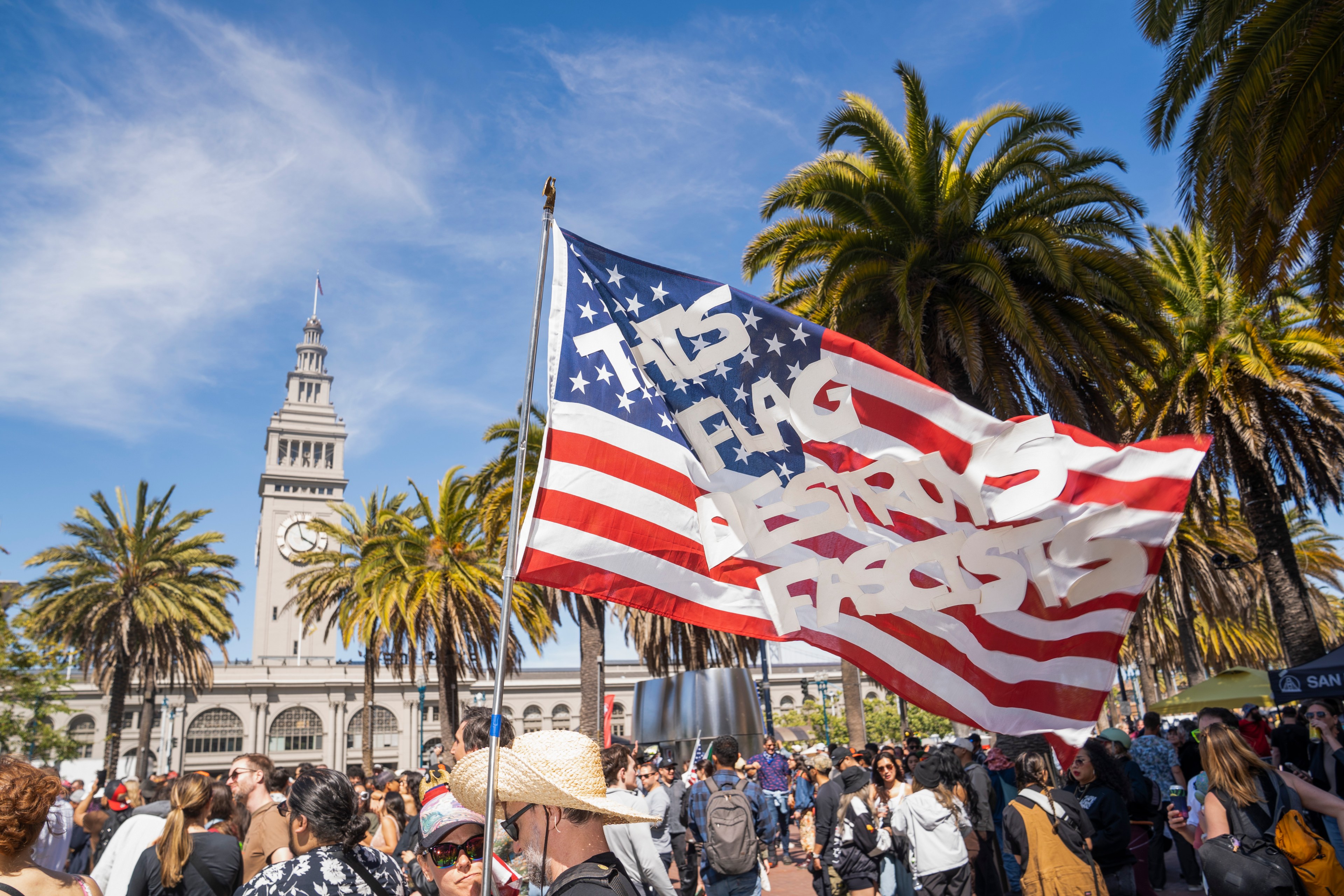 A crowd gathers near palm trees and a clock tower. An American flag with the text &quot;This flag destroys fascists&quot; is prominently displayed.