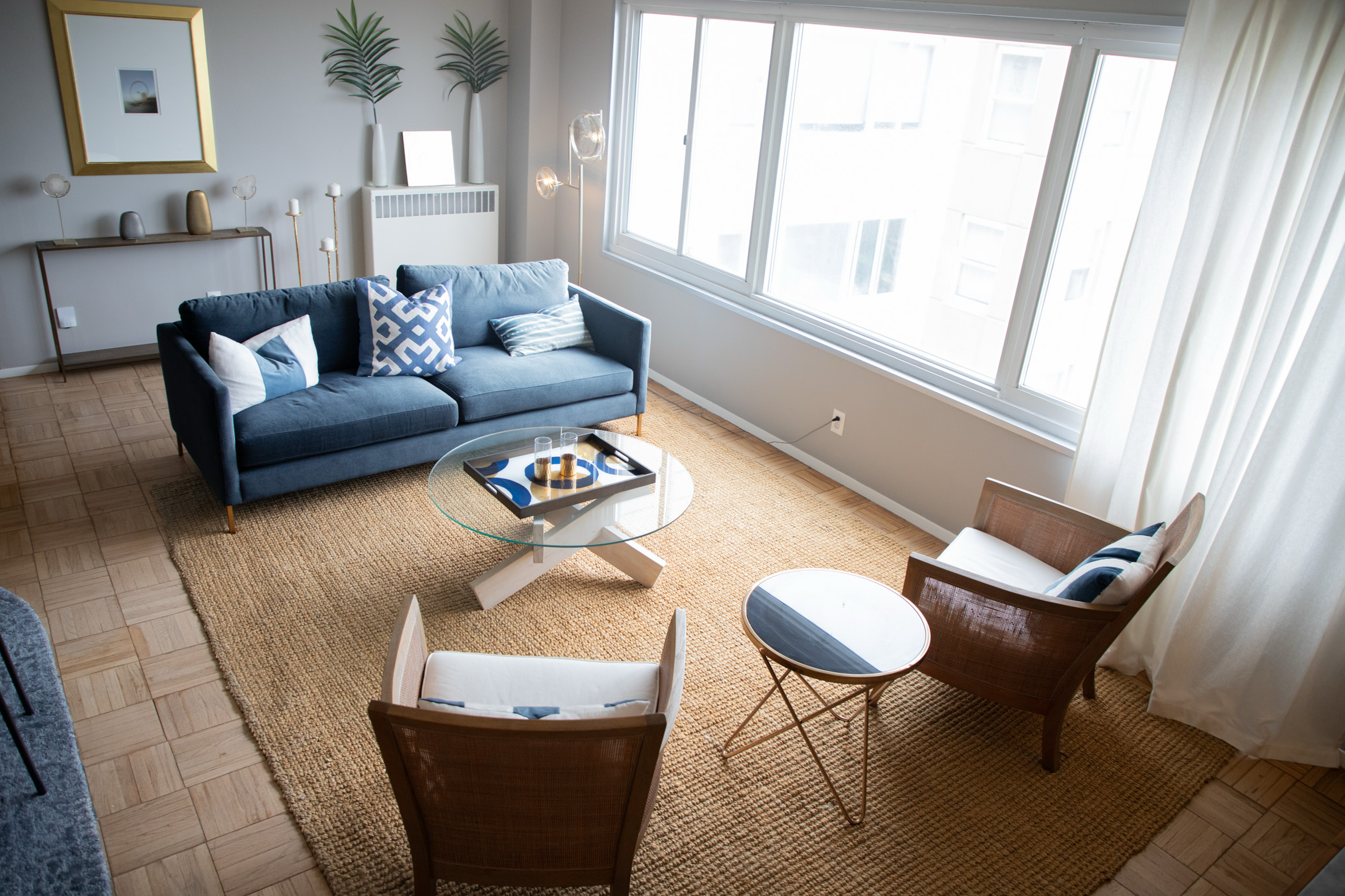 The image shows a modern living room with a blue sofa, patterned cushions, a glass coffee table, wooden chairs, and a large window with white curtains.