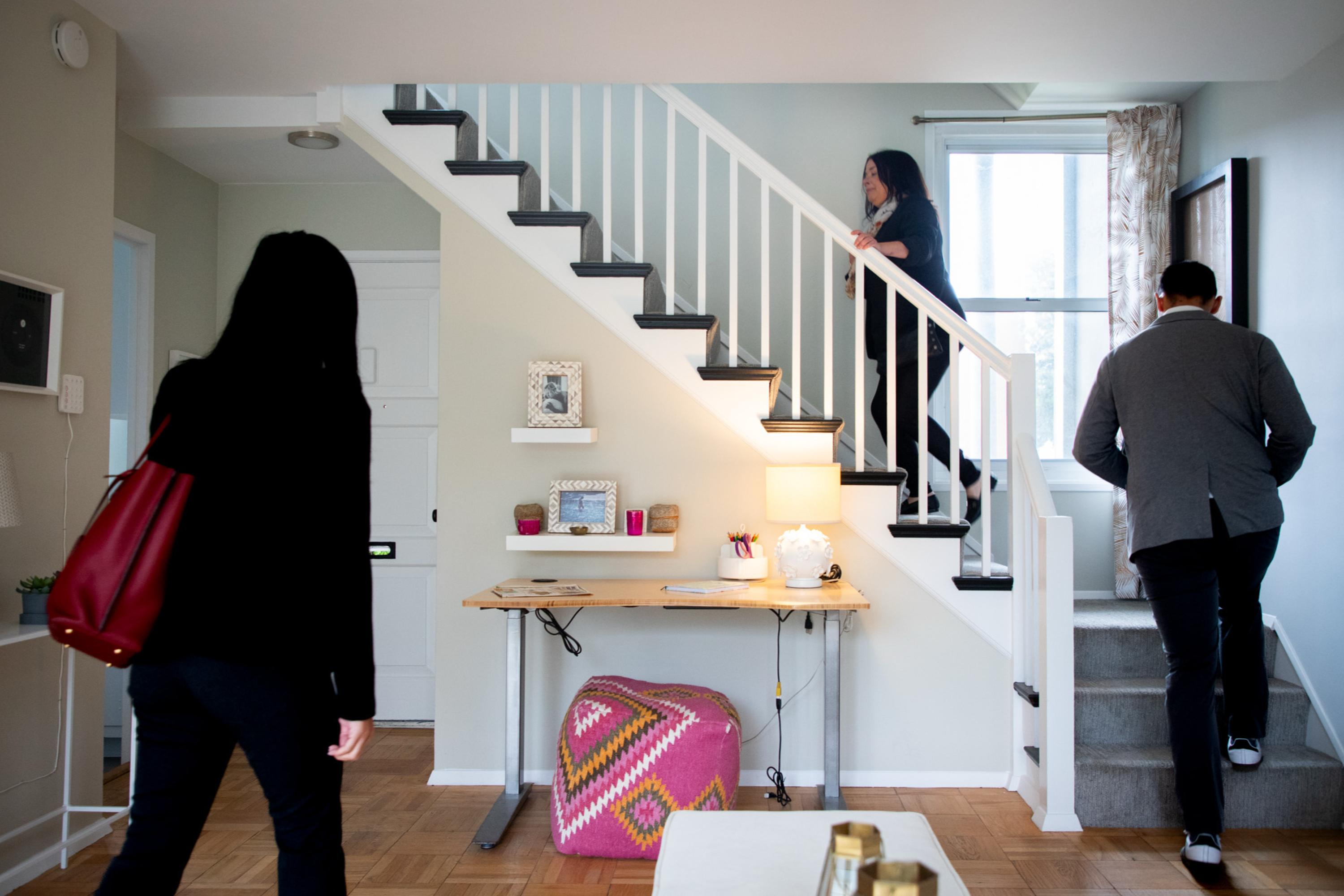 A modern living space features a staircase with two people ascending. One person is also entering. The room has a desk, shelves, and a window with natural light.
