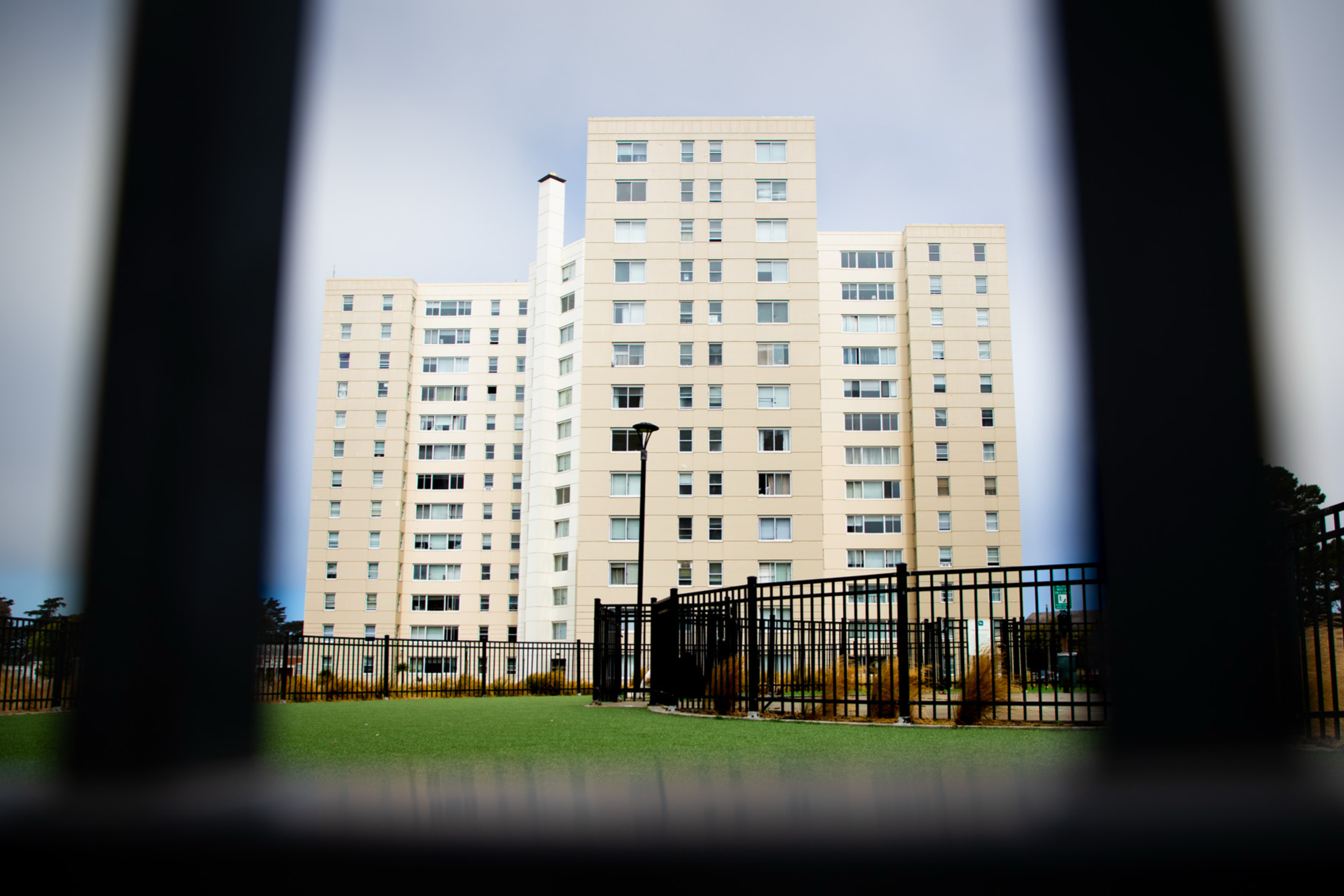 A tall, beige apartment building stands under a cloudy sky, seen through a black fence, with a green lawn and lamppost in the foreground.