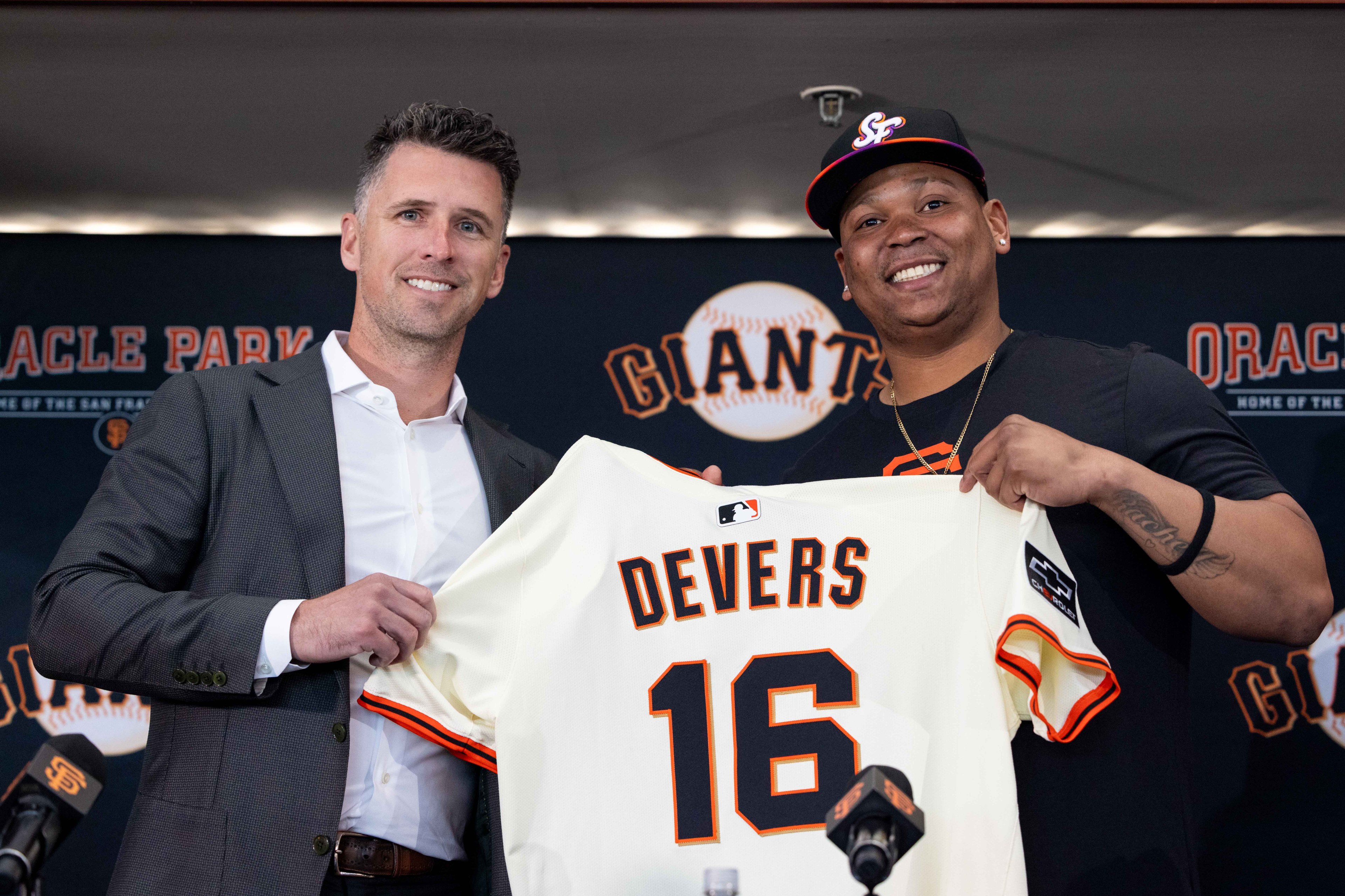 Two men are smiling on a stage, one in a suit and the other in a cap, holding up a baseball jersey that says "Devers 16" with a Giants logo in the background.