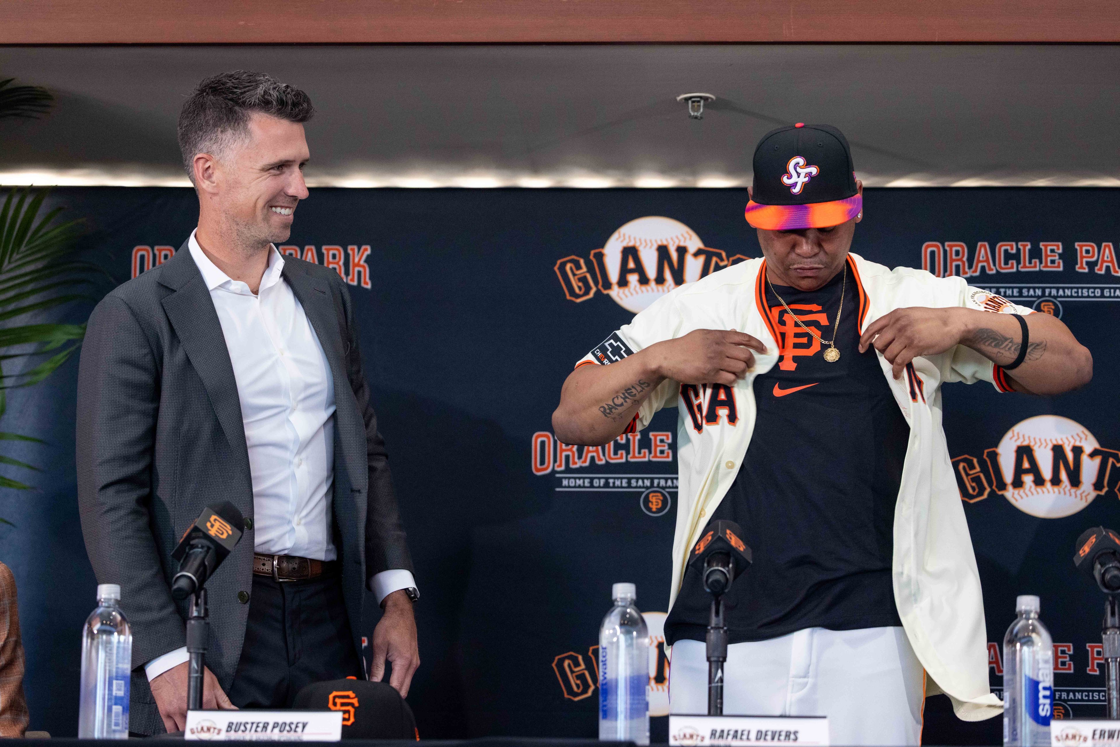 Two people stand at a podium with microphones. One is in a suit smiling, the other wears a baseball jersey and cap, adjusting his shirt. The background says "Giants" and "Oracle Park".