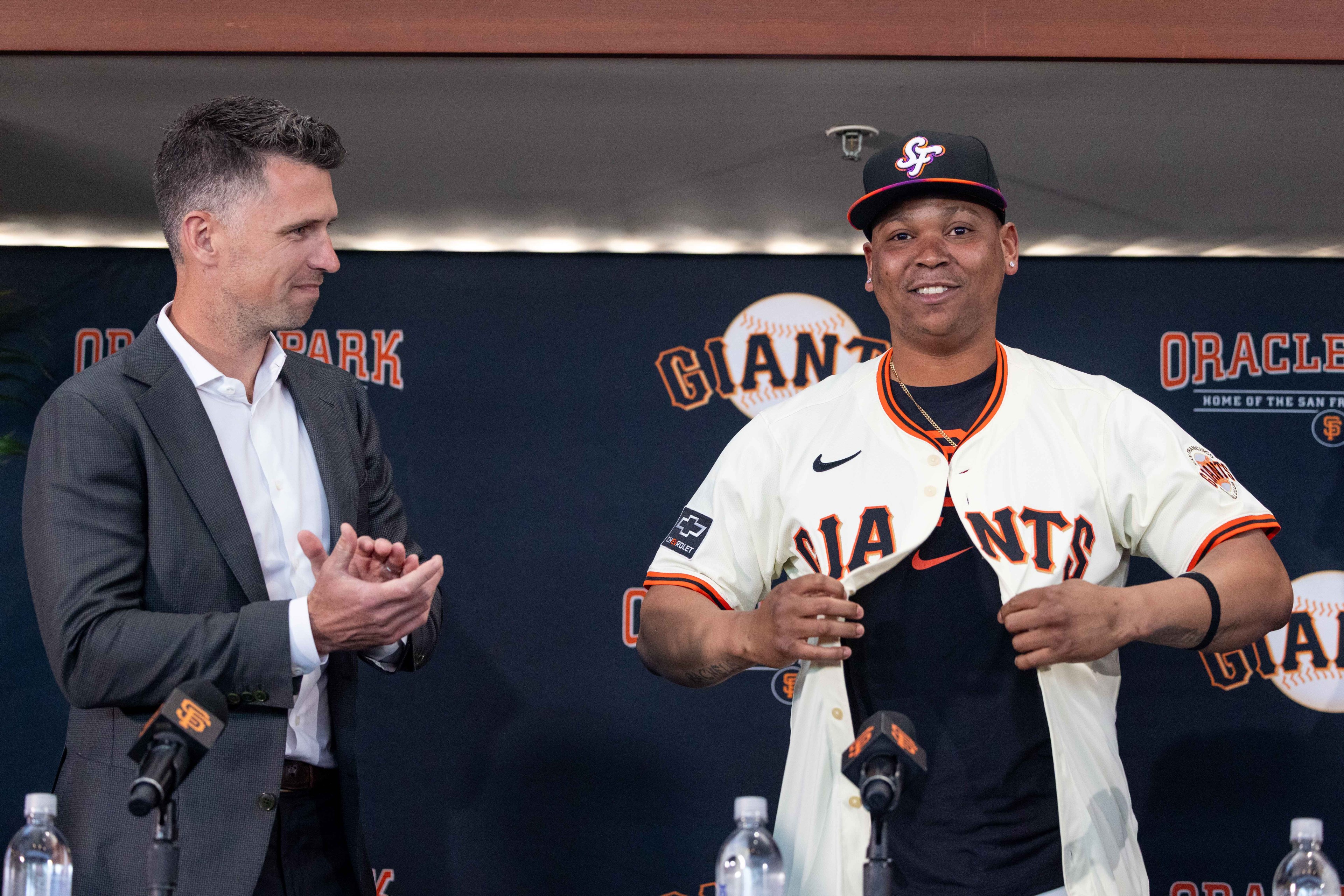 Two men are at a press event. One is clapping, while the other is smiling in a baseball uniform. Microphones and bottled water are on the table.