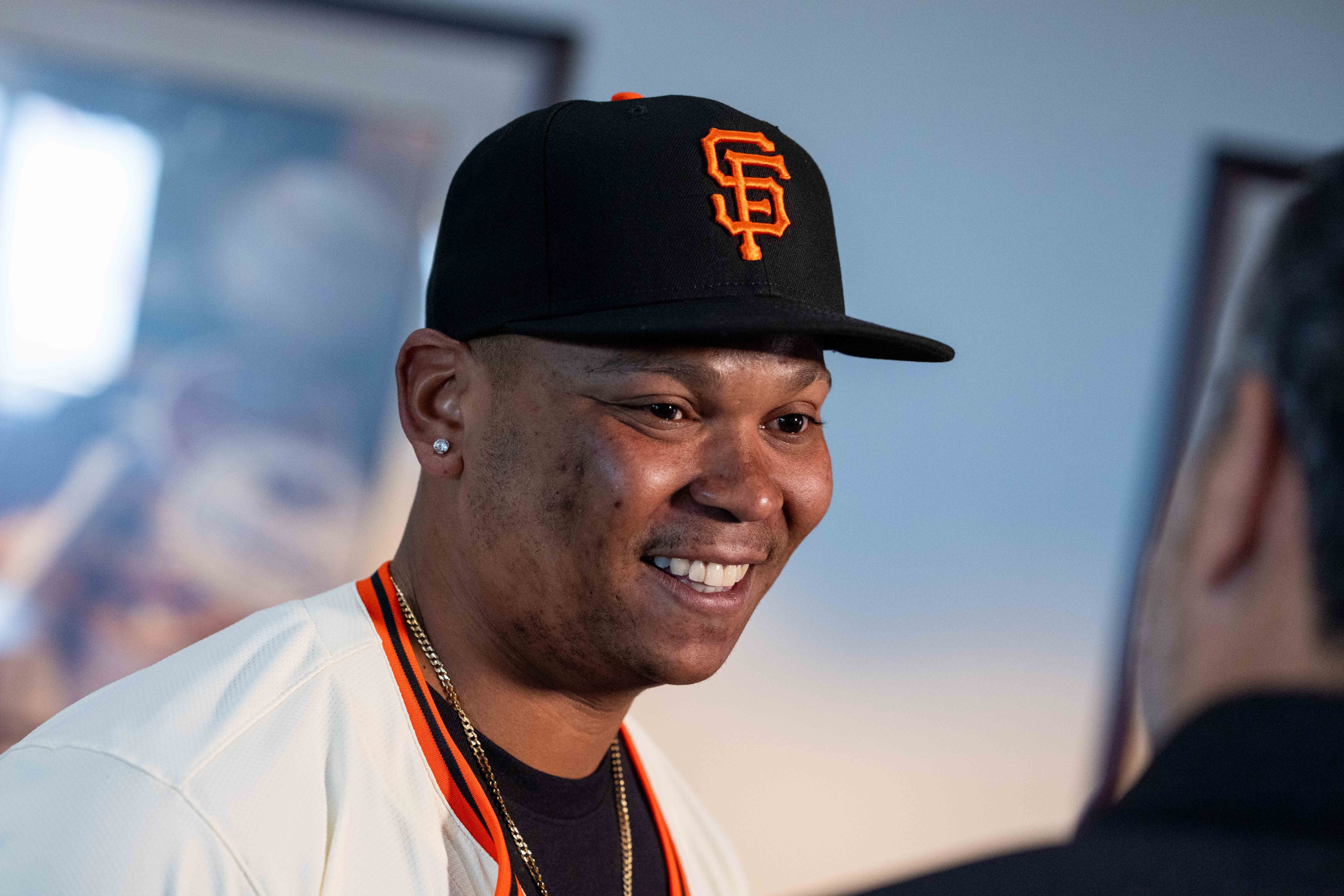 A person wearing a black cap with an orange logo smiles, facing another individual. They are dressed in a white jersey with black and orange trim.